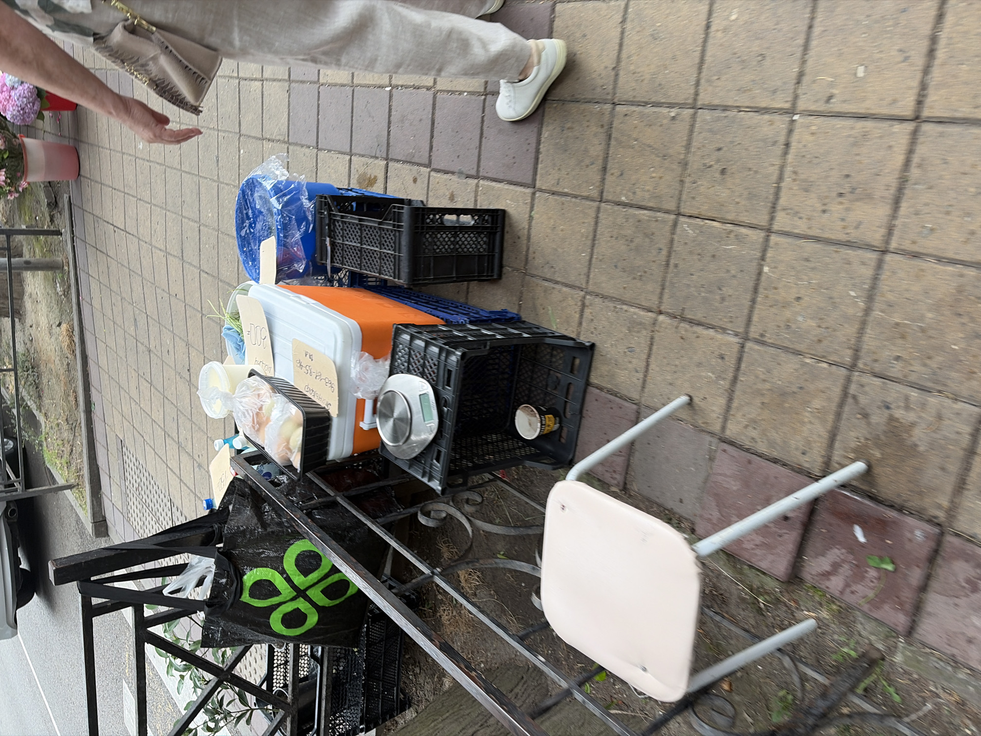 A sidewalk scene in Sochi, Russia, shows a collection of items for sale. In the foreground, a light-colored folding chair sits on a tiled surface. Behind the chair, a black metal railing encloses a collection of goods. A large black tote bag with a green clover-like logo is prominently displayed. Next to it, stacked black plastic crates hold various items. One crate contains an orange and white cooler with a clear plastic bag of bread inside. A digital scale rests on another crate. A small paper cup with some liquid is visible in a lower crate. A large blue plastic bin covered with a clear plastic bag and a blue lid is also present, along with more stacked black crates. In the background, a person's legs, wearing light-colored pants and white shoes, are visible, suggesting they are the vendor. The ground is paved with square tiles, some of which appear wet. Further in the background, a vehicle is partially visible, and potted flowers add a touch of color. The scene appears to be outdoors, on a city street or promenade.