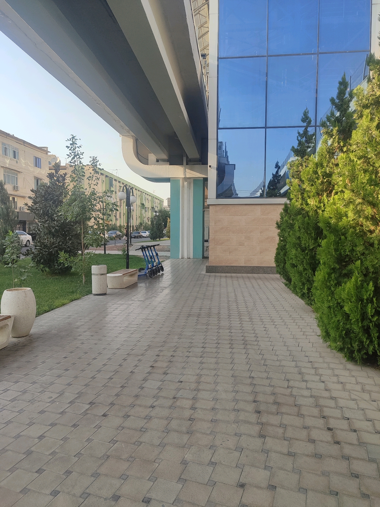 The image depicts a street scene in Tashkent, Uzbekistan, with a focus on the intersection of a modern building and an elevated roadway. A large, grey concrete overpass dominates the upper left portion of the frame, casting a shadow over a pedestrian walkway paved with grey interlocking square tiles. To the right of the walkway, a modern building features a facade of large, reflective blue glass windows. Below the windows, a section of the building is clad in light beige stone tiles.
In the mid-ground, a patch of well-maintained green grass separates the walkway from a street lined with apartment buildings. Several trees with lush green foliage are scattered throughout the scene. A row of bright green, conical evergreen trees runs along the right edge of the walkway, adjacent to the glass-fronted building.
On the pedestrian walkway, several objects are visible. A light-colored, cylindrical trash receptacle stands next to a light-colored, curved bench. To the right of the bench, four blue electric scooters are parked in a row, likely part of a rental service. In the background, cars are visible on the street, and the facade of a multi-story apartment building with numerous windows can be seen. The sky above is a clear, pale blue, indicating daytime.
