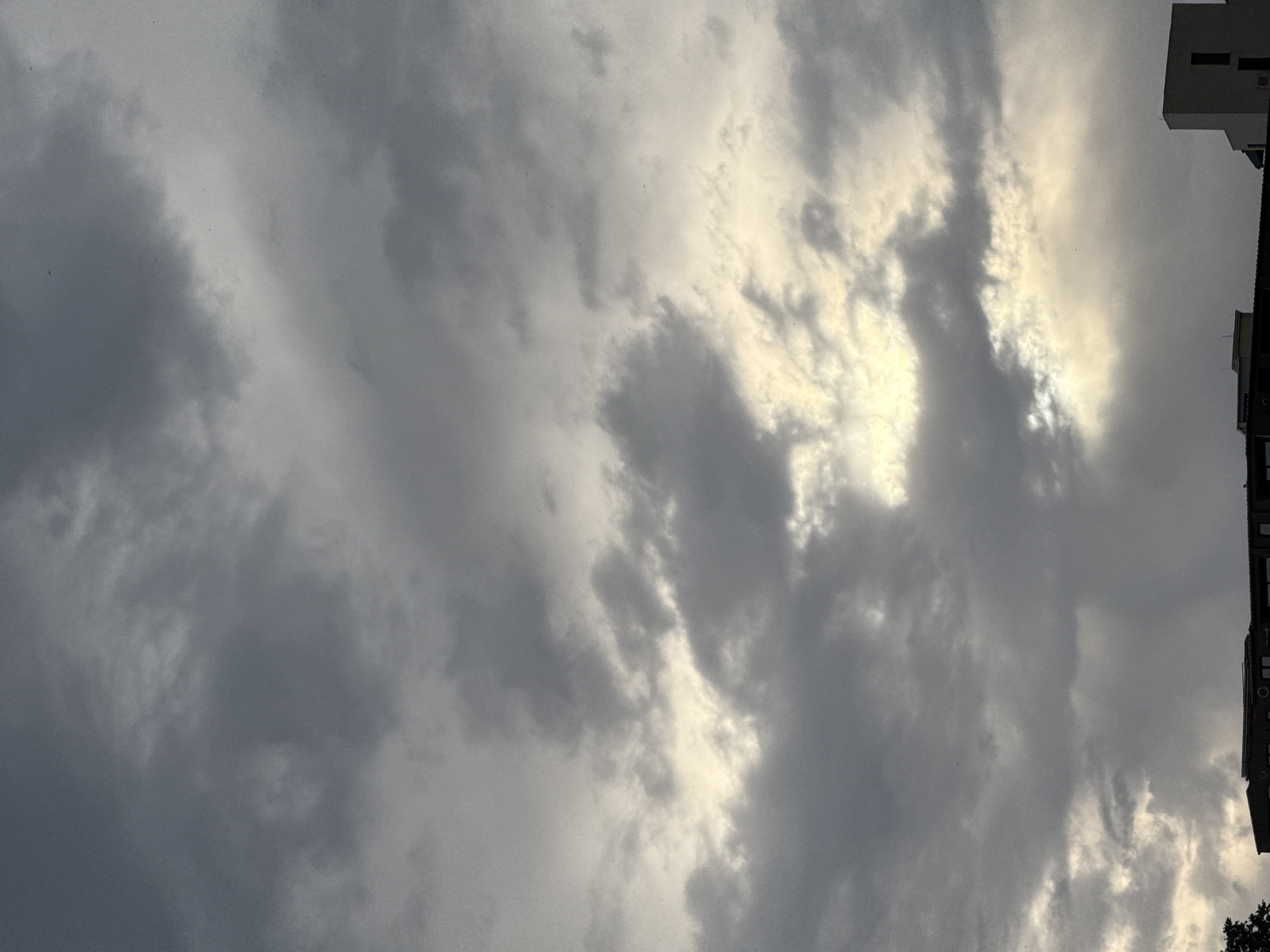 This is a photograph of a cloudy sky with buildings on the right side of the frame. The clouds are varied in texture and density, with some appearing dark and heavy, while others are lighter and more wispy. There is a bright area in the sky, suggesting the sun is attempting to break through the cloud cover. This could indicate either sunrise or sunset, but without further cues, it is difficult to determine the exact time of day. The buildings appear to be apartments or office buildings, with multiple windows visible. The overall mood of the image is somewhat dramatic due to the heavy cloud formations and the contrast between the dark clouds and the bright patches of sky. There is no visible text in the image. The location context is Boreti, Montenegro.
