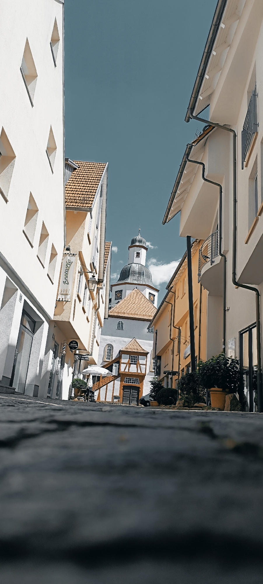 The image shows a street in a town with buildings on either side and a church tower in the distance. The street is paved with cobblestones, and there are some plants and potted trees along the side of the buildings. The buildings are a mix of modern and older architecture. On the left, there is a white building with diamond-shaped windows. On the right, there is a yellow building with a balcony. The church tower has a dark dome and a clock. The sky is blue with some clouds. There is an umbrella and chairs set up outside one of the buildings, suggesting an outdoor seating area. A black car is parked on the street. There are signs on some of the buildings, including one that says "Brasserie" and another with a barber pole image. The perspective is from a low angle, looking up towards the buildings and the sky.