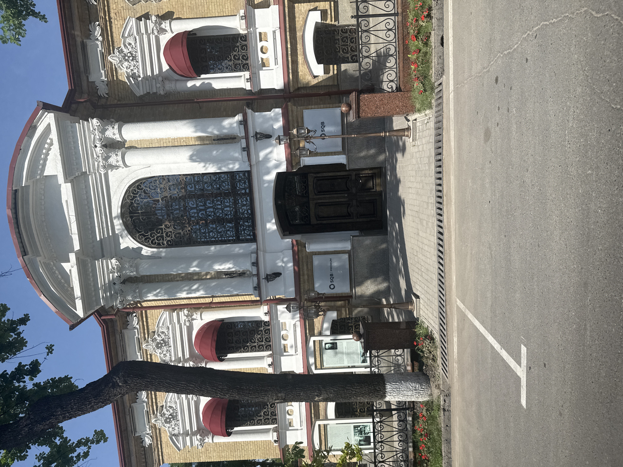 The image shows the facade of a building, likely a commercial or public institution, in Kashgar mahalla, Uzbekistan. The architecture is ornate, with white columns, intricate detailing around windows, and a rounded top section. Red awnings are above some windows. The building is constructed of light-colored brick. A dark-colored, possibly metal, door is visible at the entrance, flanked by signage for 'SQB Premium'. A decorative lamppost is to the side of the door. A tree trunk is in the foreground, partially obscuring the building. The street adjacent to the building is paved, with a white parking space marking visible. A strip of red flowers grows along the building's foundation behind a low, decorative iron fence.