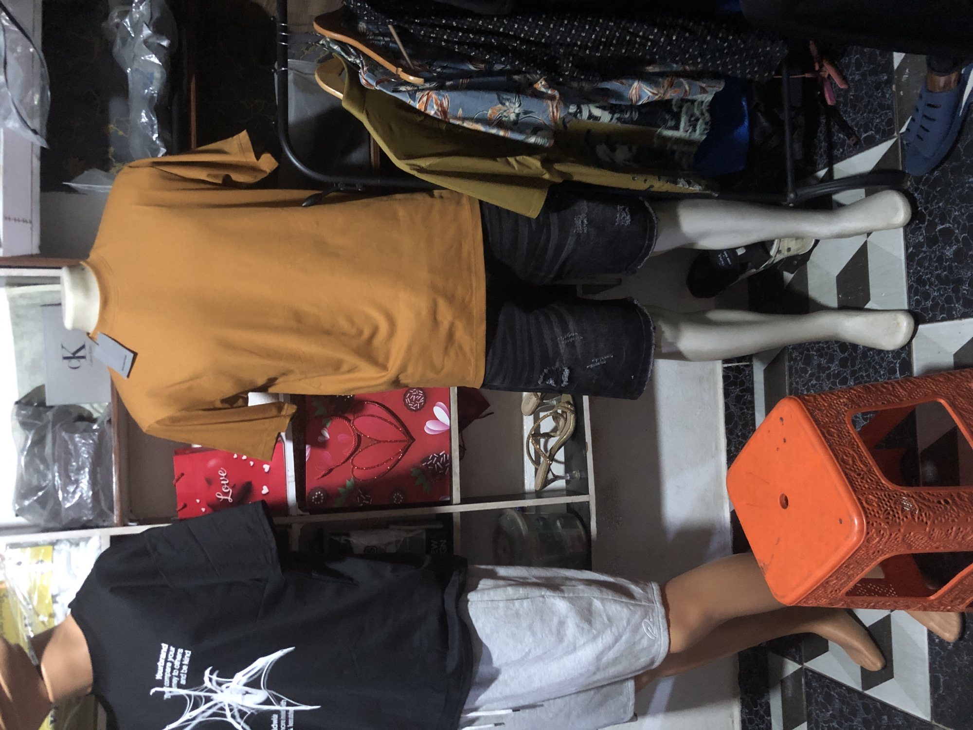 The image depicts a retail display featuring two mannequins dressed in casual clothing. The mannequin on the left wears a black t-shirt with a white graphic print and light gray shorts. The mannequin on the right is dressed in a mustard-colored t-shirt and dark denim shorts. Both mannequins are standing. In the background, shelves are visible, holding various items including shopping bags, a pair of women's sandals, and other clothing. There are also clothes on hangers on a rack. An orange stool is placed in the foreground. The flooring is a black and white tiled pattern. The scene appears to be a clothing store or a section of a market stall. The overall impression is one of a retail environment. The presence of a blue sandal on the floor suggests ongoing retail activity. The lighting is somewhat dim, creating shadows and highlighting the displayed clothing. The image is taken from a frontal perspective, at a medium distance.