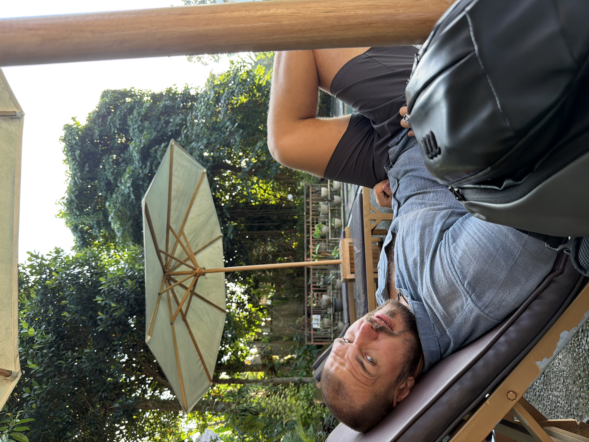 In Ubud District, Indonesia, a man with a buzz cut and beard, wearing a light blue button-up shirt and dark shorts, reclines on a brown chaise lounge. He is positioned to his right, with his head turned towards the camera and his right leg bent. A black backpack rests on his chest and lap. The chaise lounge is situated outdoors, under a light green umbrella. Behind the man, there are trees and foliage, suggesting a tropical environment. Other lounge chairs, a table, and potted plants are visible in the background. The lighting indicates it is daytime, with sunlight filtering through the trees.