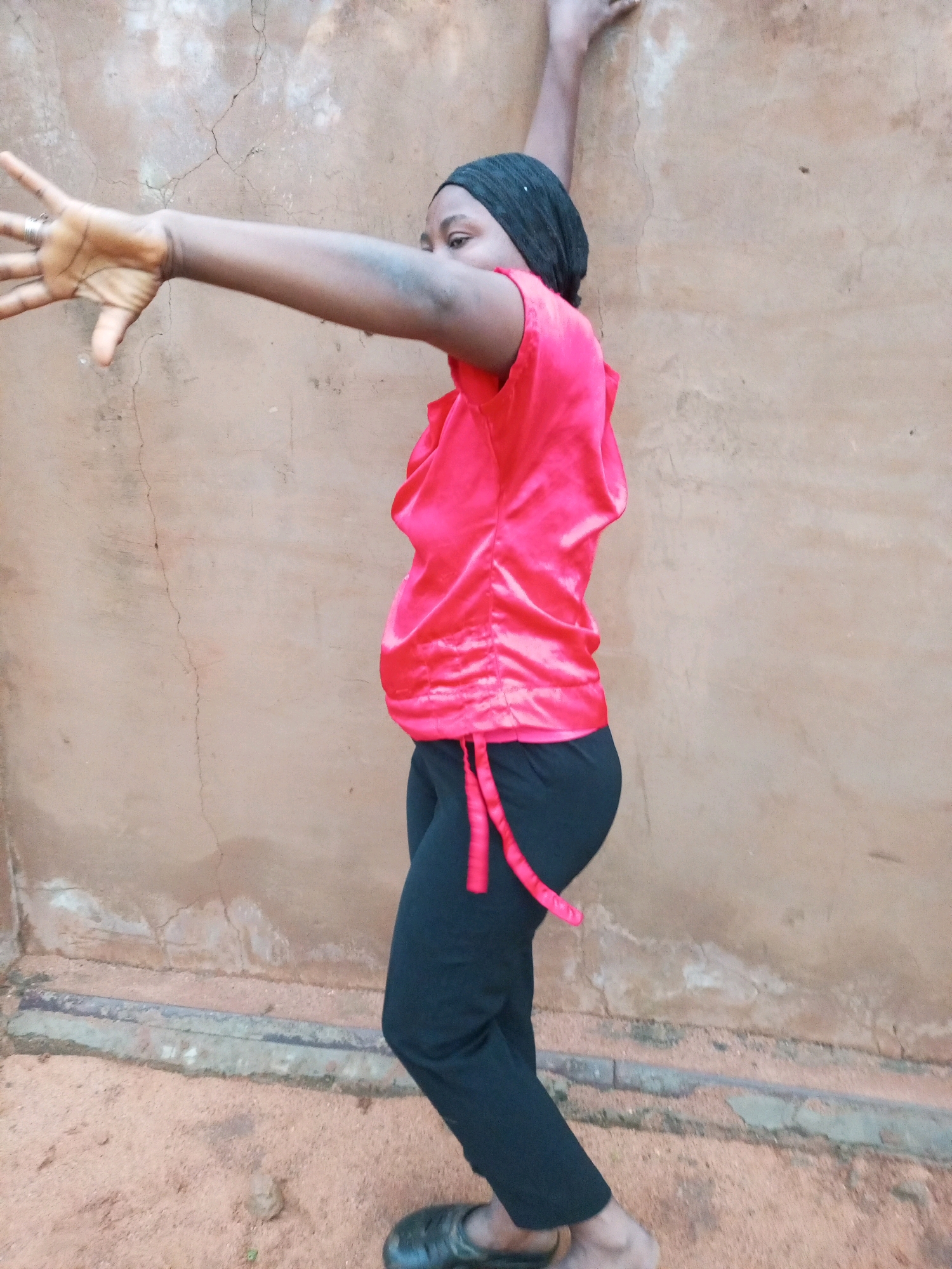 A person wearing a red top and black pants stands with their arms outstretched against a textured, light brown wall in Tempure, Nigeria. The person's left arm is extended towards the left, with their fingers splayed. Their right arm is bent at the elbow, and their hand is resting on the wall above their head. The person is wearing a black headscarf and black slip-on shoes. The ground is covered in reddish-brown sand and has a concrete gutter running along the bottom of the wall. The wall itself appears weathered, with some cracks and patches of discoloration.
