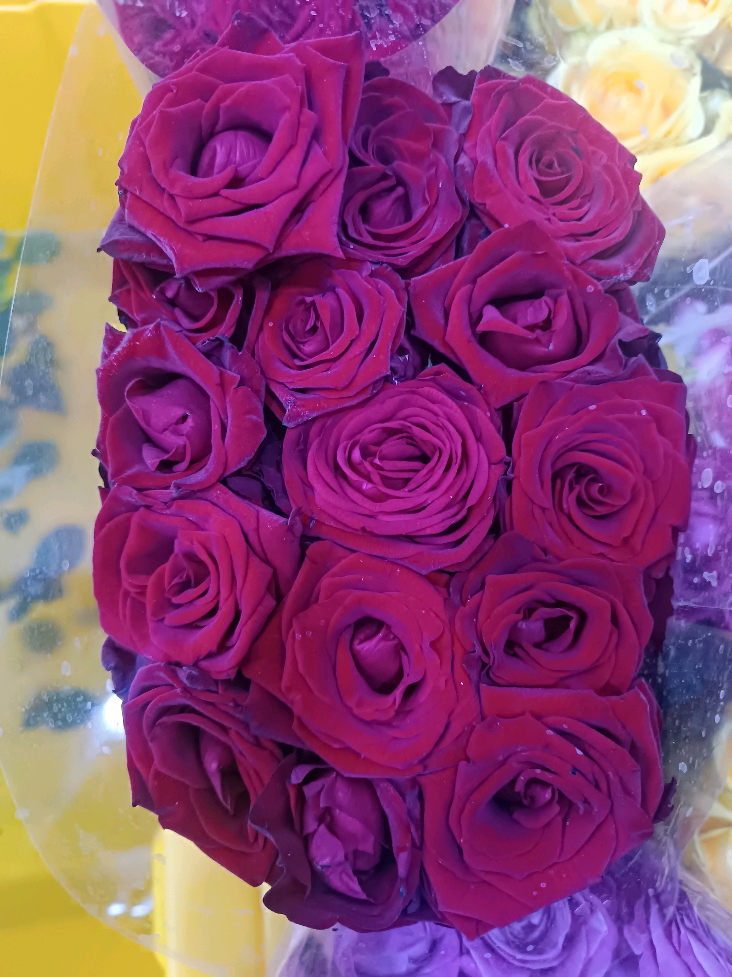 A bouquet of deep red roses is presented in a plastic wrap. The roses are tightly packed together, with some of them showing water droplets on their petals. In the background, out of focus, are yellow roses and a vibrant yellow surface, possibly a stall or display. The overall impression is that of a flower shop or market in Yangiyer, Uzbekistan, where the flowers are arranged for sale.