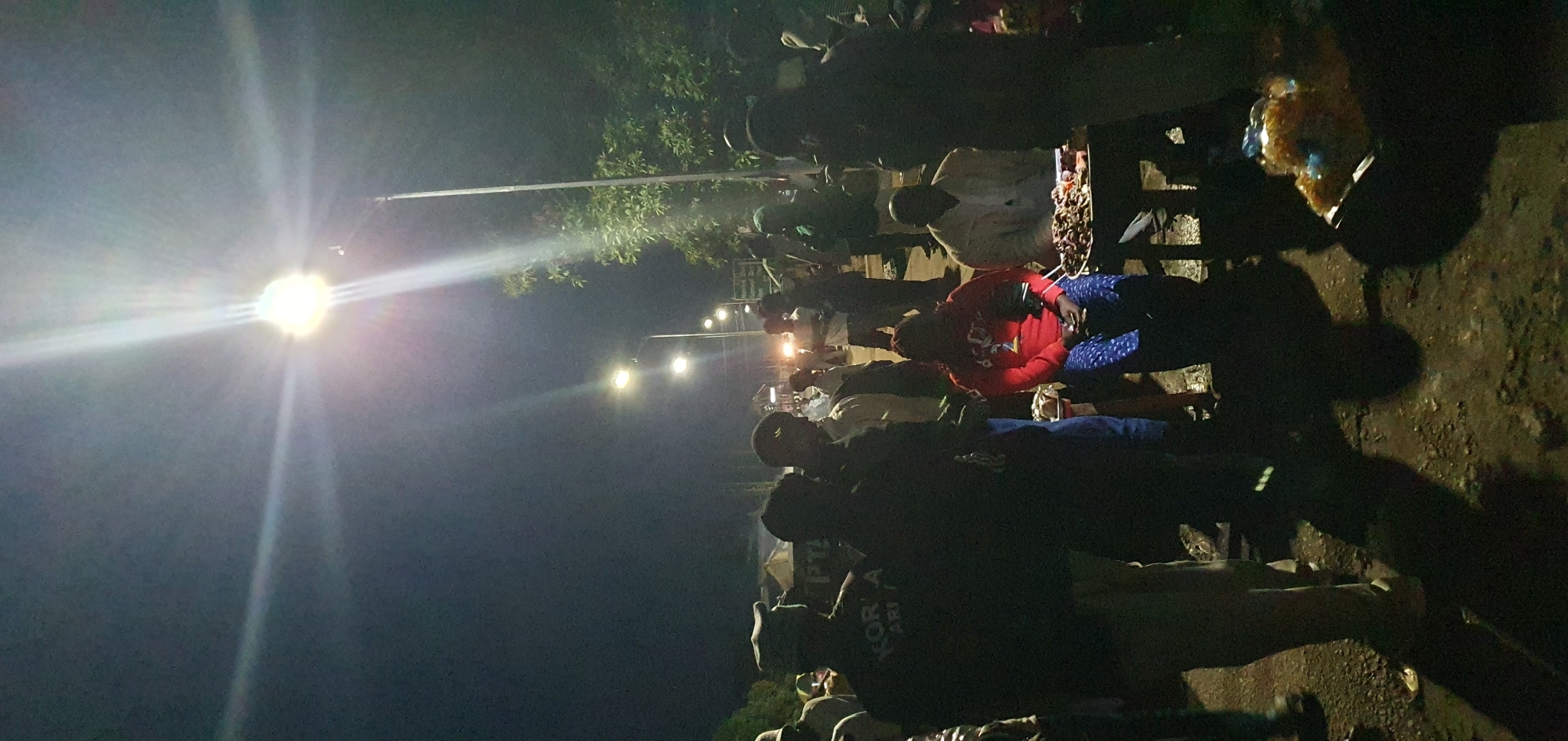 A nighttime outdoor scene in Bukuru, Nigeria, is illuminated by bright streetlights, creating stark shadows. A group of people are gathered on a dirt surface, some standing and others seated. In the foreground, on the left, a person in a dark jacket with white lettering on the back is visible. To the right of them, a person in a red top and blue patterned pants is seated near a small table with goods. Another individual is seated to the right of the person in red, wearing a light-colored jacket. Several other individuals, some appearing to be men in dark clothing and others in lighter attire, are dispersed throughout the scene. A table laden with food items is positioned in front of the seated individuals. In the background, streetlights line a road, and the headlights of vehicles are visible. Trees are present on the right side of the image, and some tents or structures are visible in the distance on the left. The ground is uneven and appears to be a mixture of dirt and gravel.