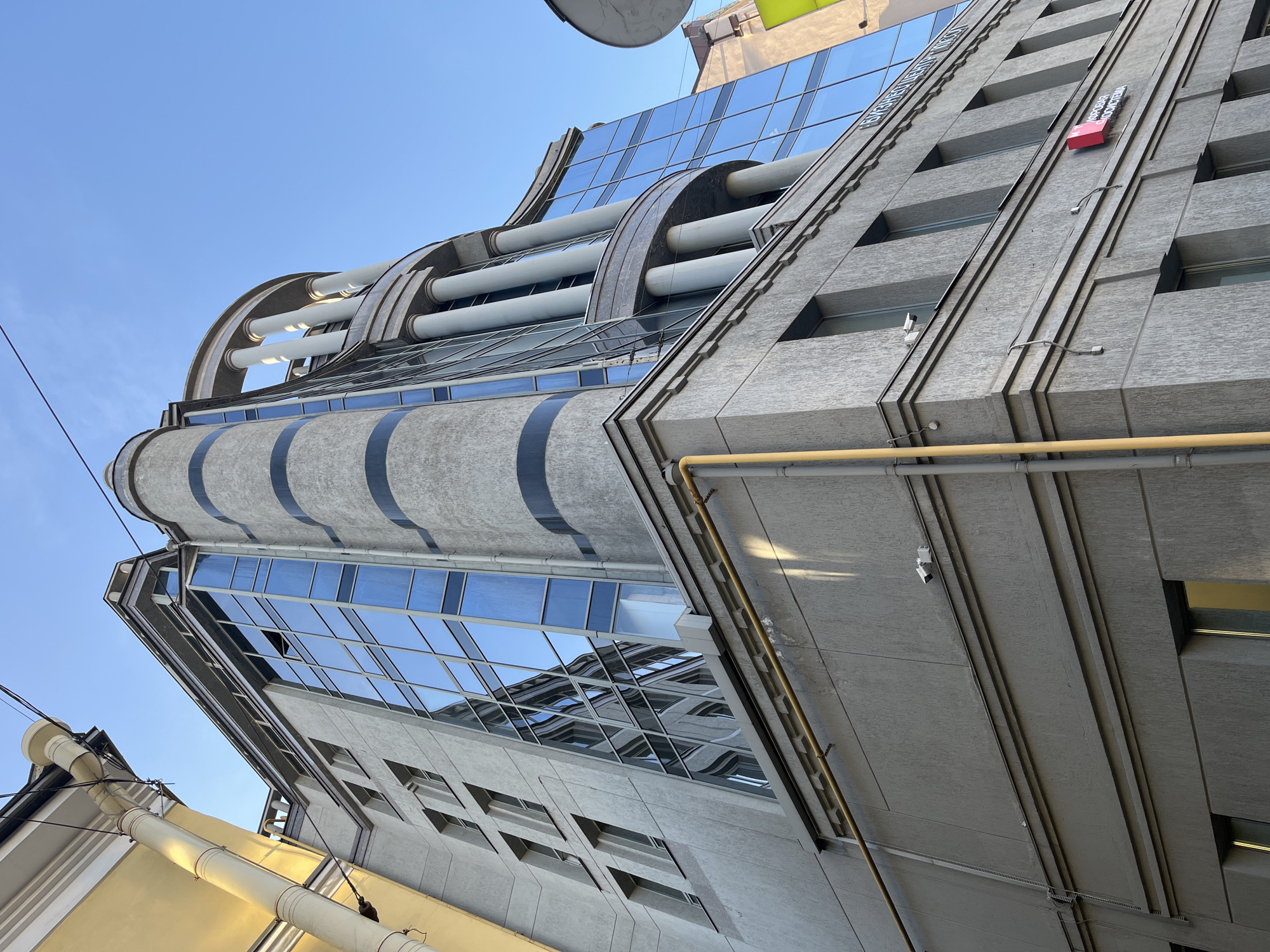 The image shows a low-angle view of a modern building in Saint Petersburg, Russia. The building features a combination of concrete and glass facade. The concrete sections have deeply recessed windows. The glass portion of the building has a curved design at the top, supported by cylindrical structures. There are yellow pipes running along the concrete section of the building. A red box with Russian text is mounted on the wall. Cables are visible in the foreground. The sky is clear and blue.