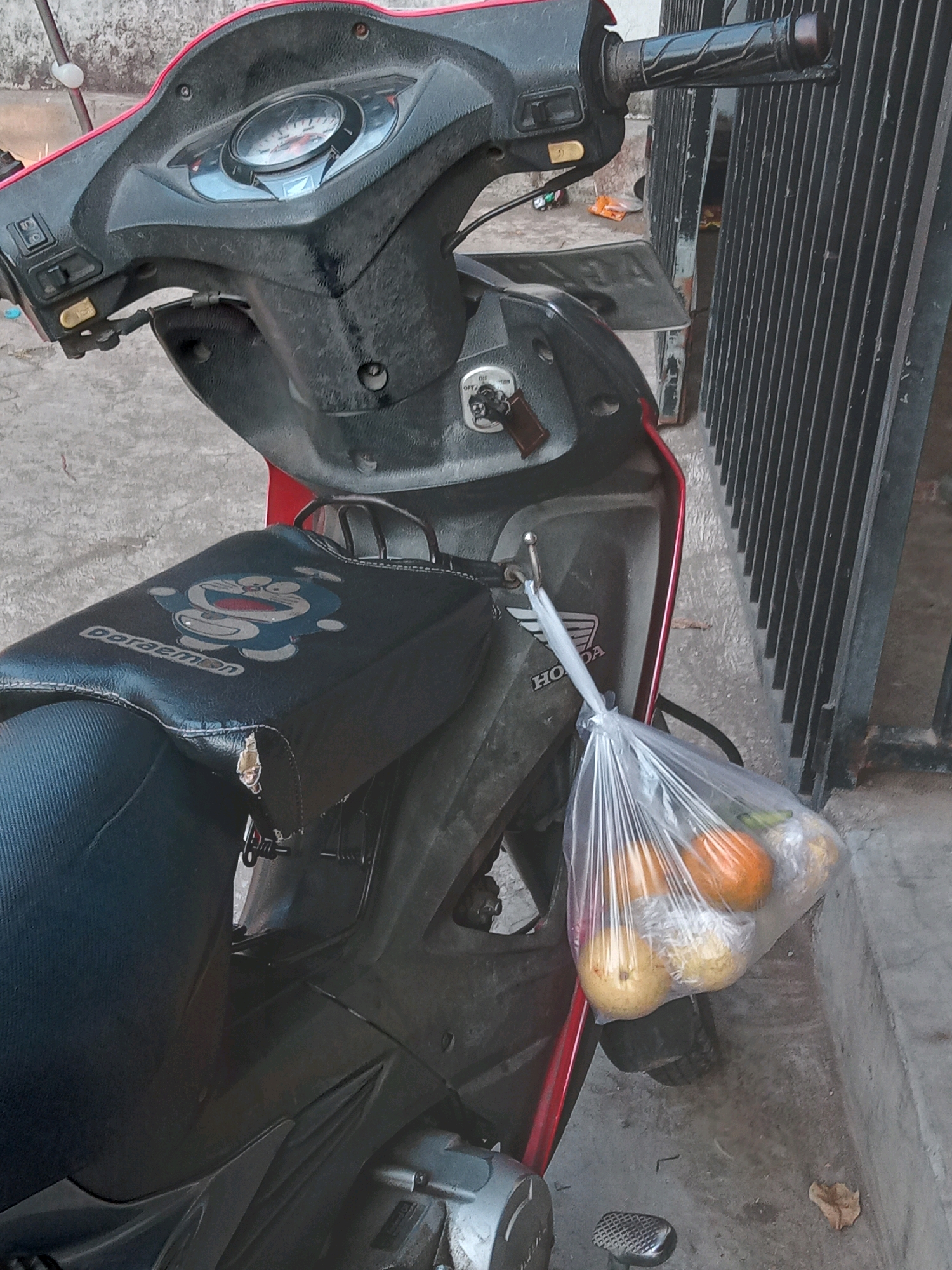 This is a medium-shot, eye-level, close-up photograph taken outdoors in Sukoanyar, Indonesia. The primary subject is a black and red motorcycle. The motorcycle has a seat cover with a depiction of the cartoon character Doraemon. A clear plastic bag containing several pieces of fruit, including what appear to be two yellow guavas and two orange fruits, hangs from a hook on the side of the motorcycle. The background shows a gray concrete ground and a black metal gate. There are no people visible in the image. The lighting suggests it is daytime, with some shadows present. The overall scene appears to be a street or sidewalk area.