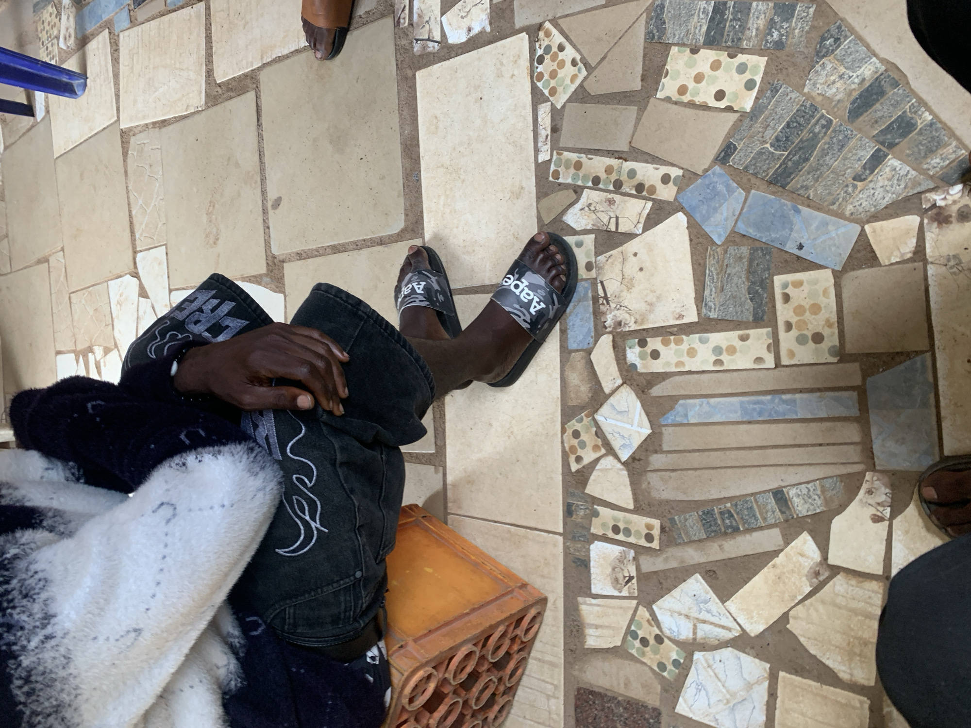 In Bukuru, Nigeria, a close-up shot captures a person seated on an orange plastic crate with a patterned tiled floor. The individual wears a fuzzy, black and white patterned sweater and dark denim jeans with white stitching. On their feet are black slide sandals with a camouflage pattern and the word "Aape" printed on them. To the right, another person's foot is visible, wearing a brown sandal. The floor is a mosaic of various sized tiles, predominantly in earthy tones with some blue and patterned pieces. The background is out of focus, but suggests an indoor or semi-covered outdoor setting with a blue chair leg visible at the top right.