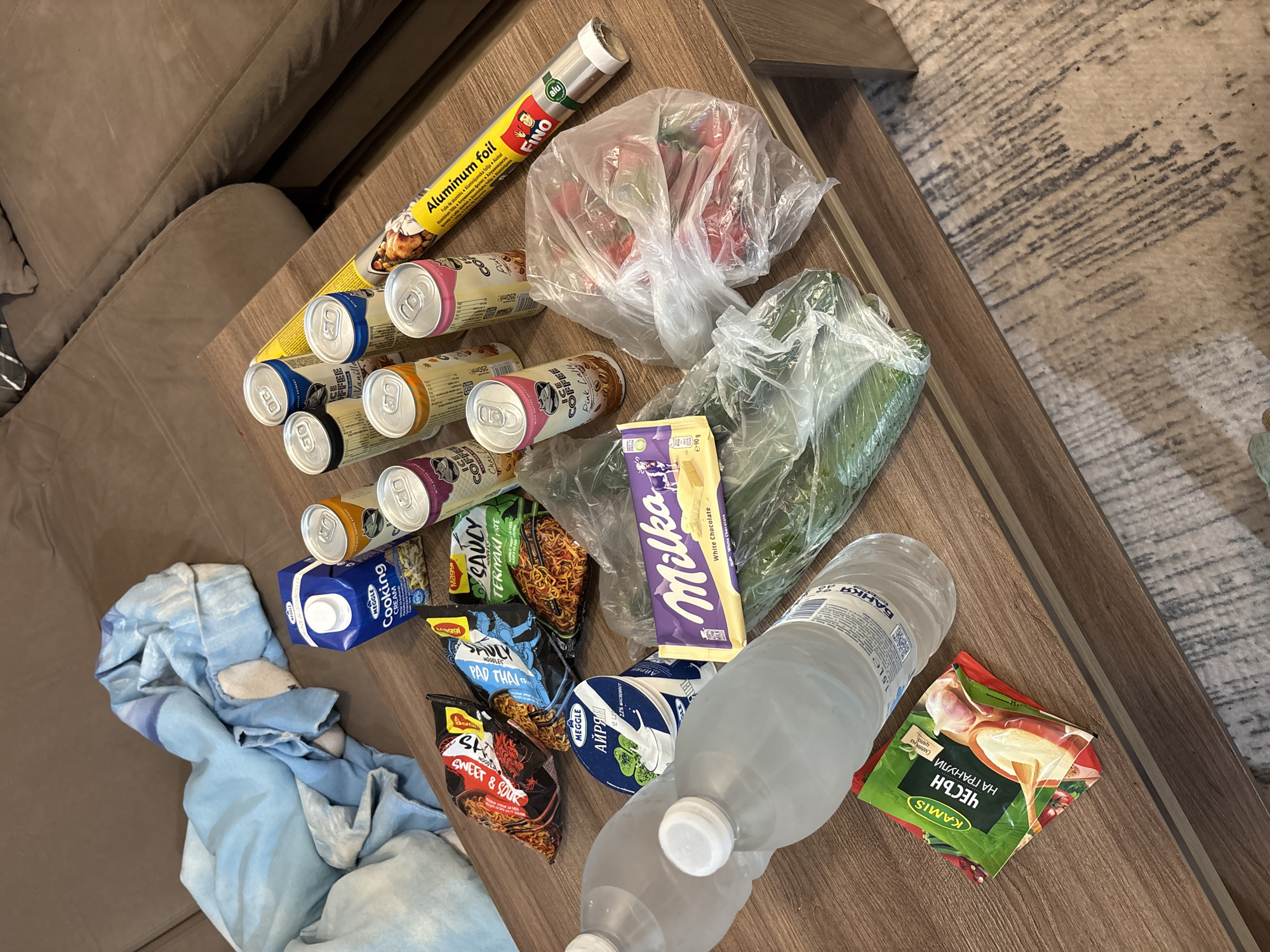 This media file depicts an assortment of recently purchased groceries laid out on a light brown wooden table, suggesting an indoor setting like a living room or dining area in Plovdiv, Bulgaria. To the left, a portion of a brown sofa is visible, and to the right, a light-colored patterned rug. A crumpled blue and white cloth rests on the sofa.
The grocery items include several cans of "ICE COFFEE" in various flavors such as Vanilla, Chocolate, and Pink Latte, some labeled as "LACTOSE FREE". There's a roll of "FINO Aluminum foil" and two clear plastic bags containing fresh vegetables, appearing to be tomatoes/peppers and cucumbers. Other items include a "Milka White Chocolate" bar (90g), multiple packages of Maggi instant noodles ("SAUCY TERIYAKI TASTE," "PAD THAI," and "SWEET & SOUR"), a carton of "MEGGLE Cooking CREAM," and a container of "MEGGLE АЙРЯН" (Ayran) with "2.7% МАСЛЕНОСТ" (fat). A packet of "KAMIS ЧЕСЪН НА ГРАНУЛИ" (Garlic Granules) and two 1.5-liter bottles of "БАНКЯ" (Bankya) mineral water are also present.
The image captures the aftermath of a shopping trip, with various food and household items awaiting storage. The presence of Bulgarian brand names and Cyrillic script, along with the provided context, confirms the location. The arrangement of items indicates they have just been placed on the table, possibly for organization or preparation.