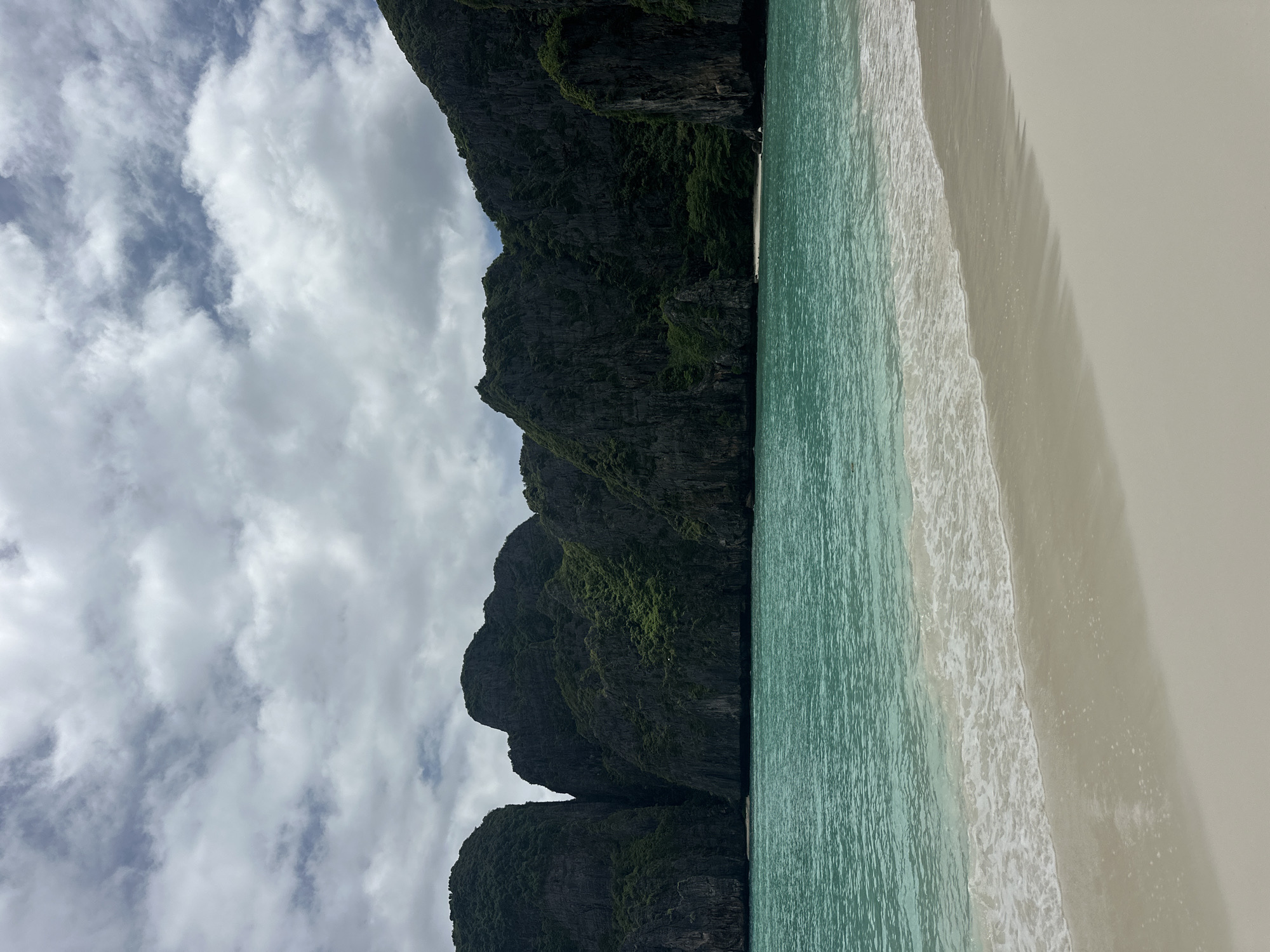 This is an eye-level, medium shot of a tropical beach scene in Ao Nang, Thailand. The foreground features a pristine, white sand beach with gentle waves lapping at the shore. The turquoise water is calm and clear, transitioning to a deeper blue as it recedes. In the background, towering limestone karst cliffs, covered in lush green vegetation, rise dramatically from the sea. The sky is overcast with scattered clouds, suggesting a moderately bright, diffused light. There are no people or man-made objects visible in the scene. The overall impression is one of natural beauty and tranquility.