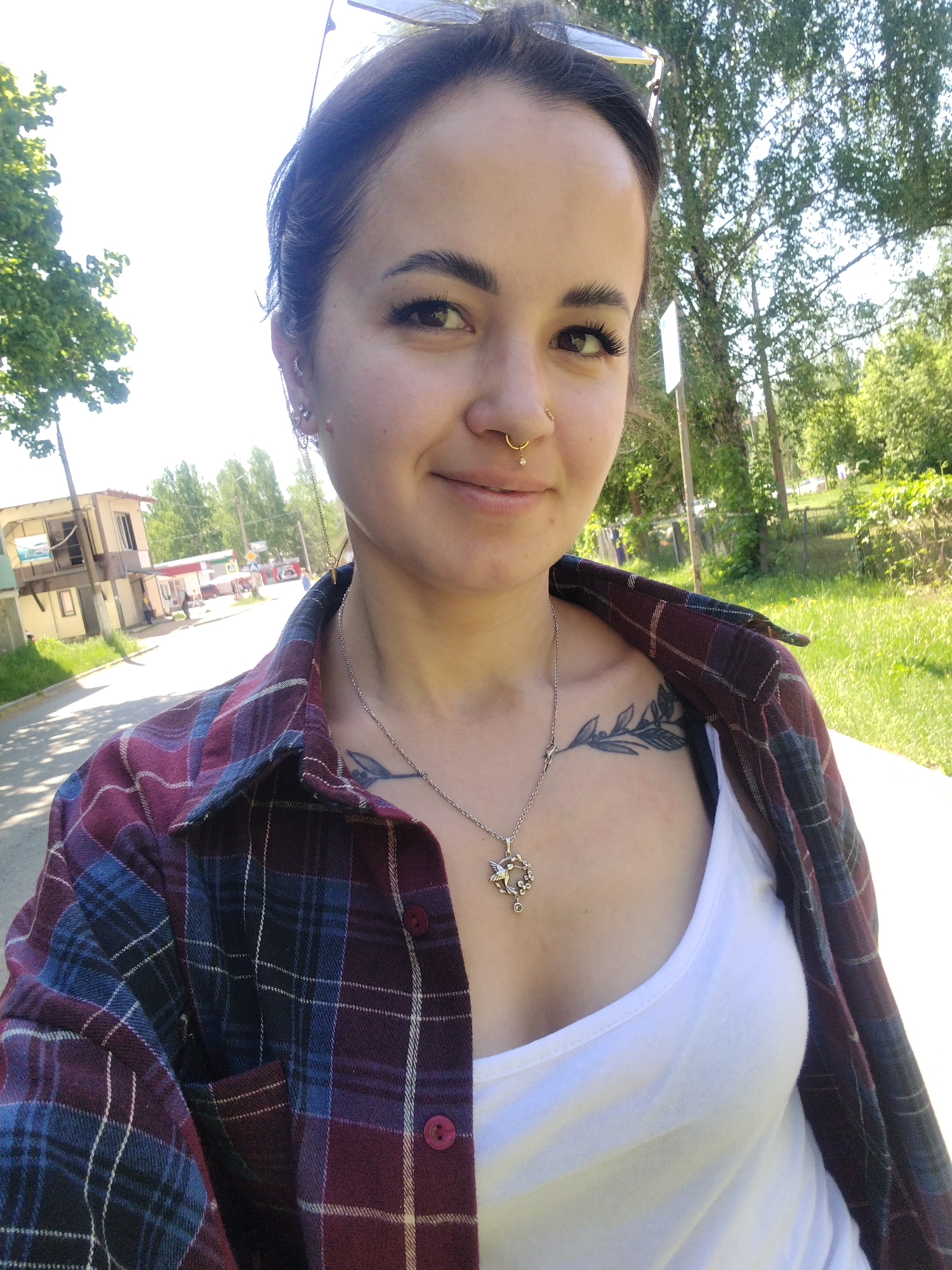 A medium shot depicts a woman in a casual outdoor setting, seemingly on a city street in Noviy, Russia. She wears a white tank top under an unbuttoned plaid shirt in shades of burgundy, blue, and black. A silver necklace with a hummingbird pendant adorns her neck, just above a tattoo on her upper chest depicting a leafy branch. The woman has dark hair pulled back, and her features include a nose ring and multiple earrings. In the background, a paved street stretches into the distance, lined with buildings, trees, and some parked vehicles. The overall environment suggests a sunny day in a suburban or residential area.