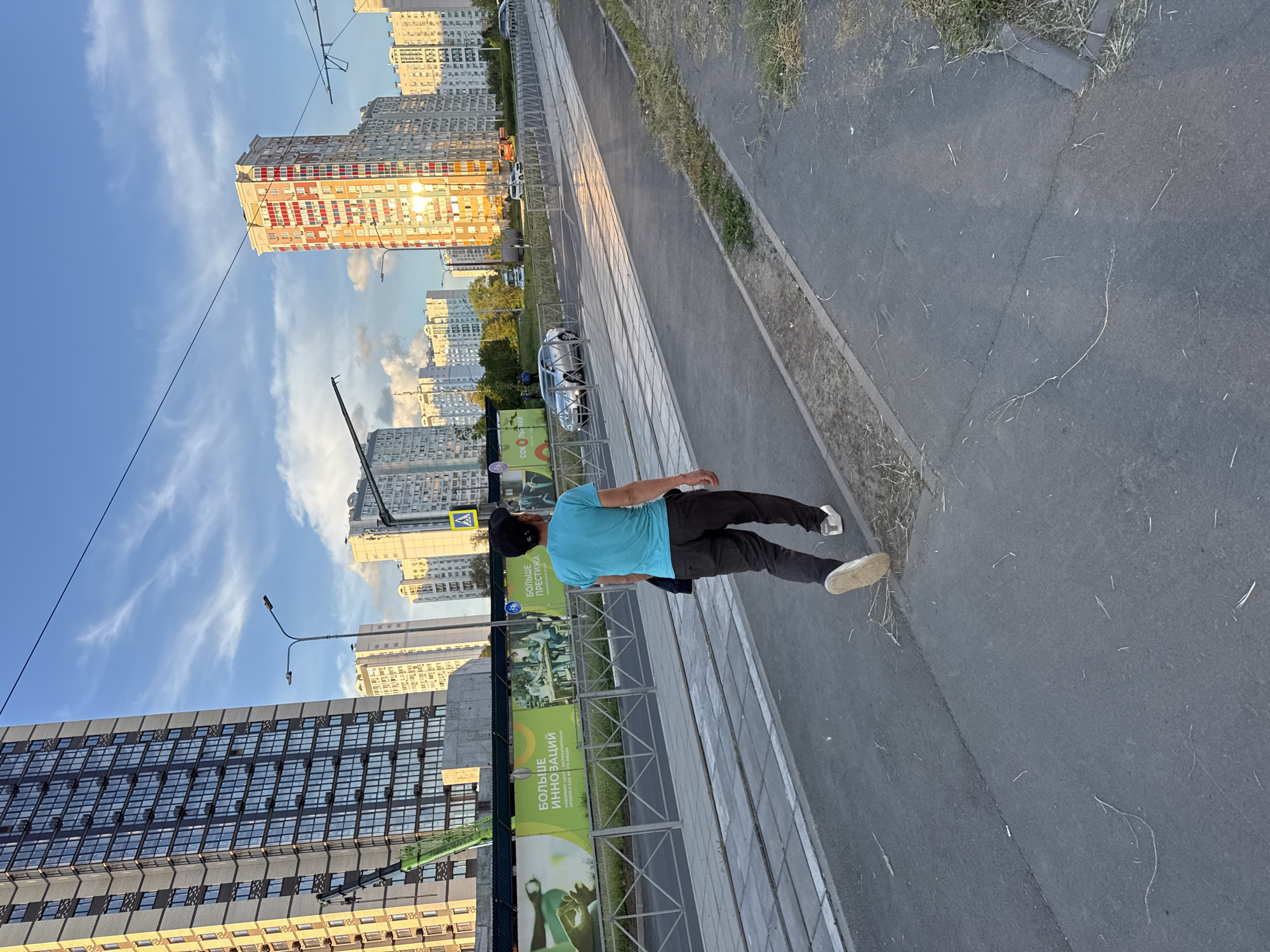 A street-level, eye-level shot shows a man walking on a sidewalk in Chaltyr', Russia. He is wearing a blue shirt, dark pants, and light-colored sneakers. His back is to the camera. A construction site borders the sidewalk with green and white promotional banners along a metal fence. High-rise apartment buildings are visible across the street under a blue, cloudy sky. The sidewalk is gray and abuts a narrow strip of vegetation. A car is parked further down the street.