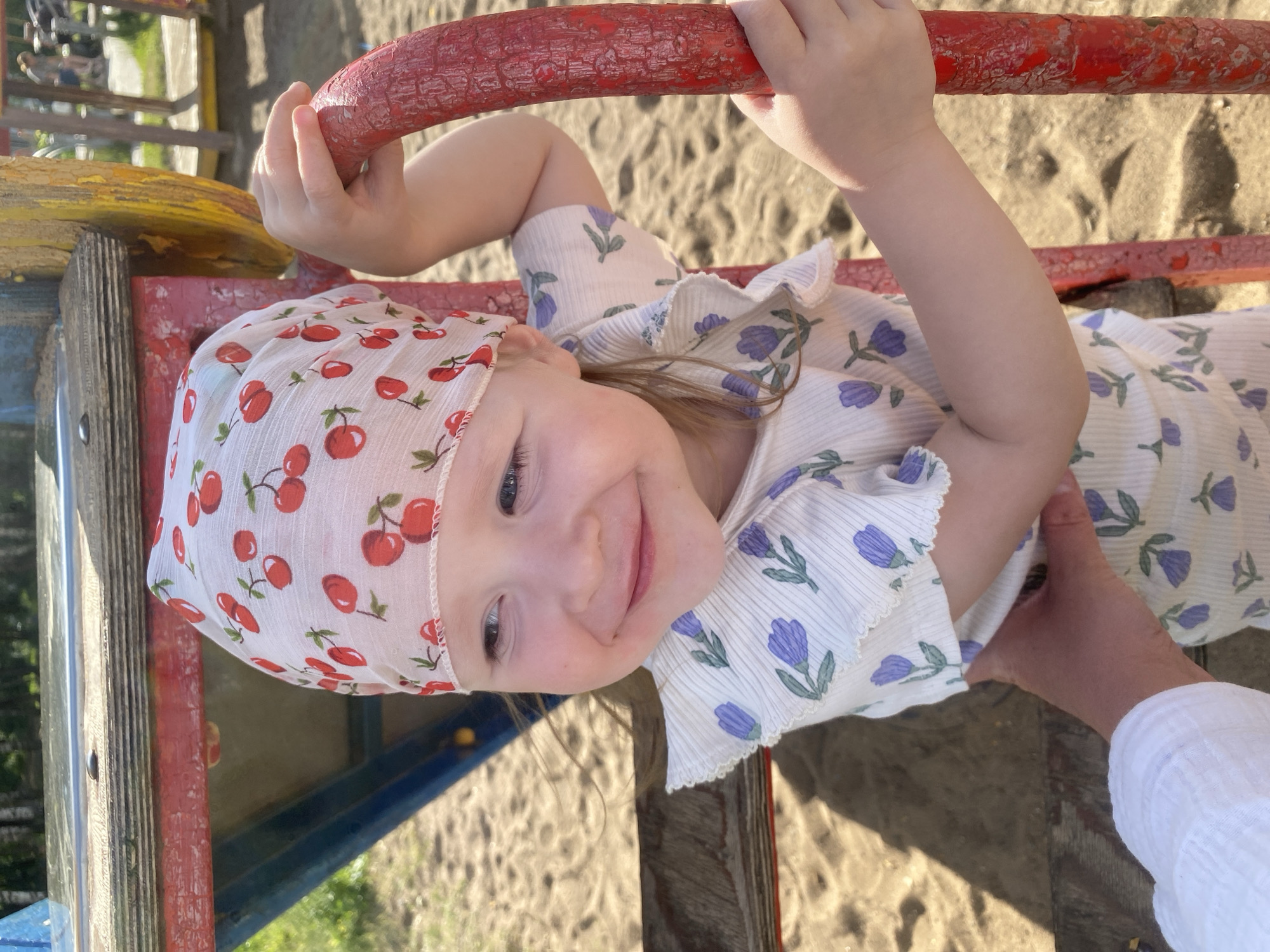 In an outdoor playground scene, a young Caucasian girl with blonde hair, wearing a white bandana with a cherry pattern and a white dress with a purple floral print, smiles as she holds onto a red, textured bar. The background shows sand and parts of a colorful playground structure. The lighting suggests a sunny day. The location is identified as Chaikovsky, Russia.
