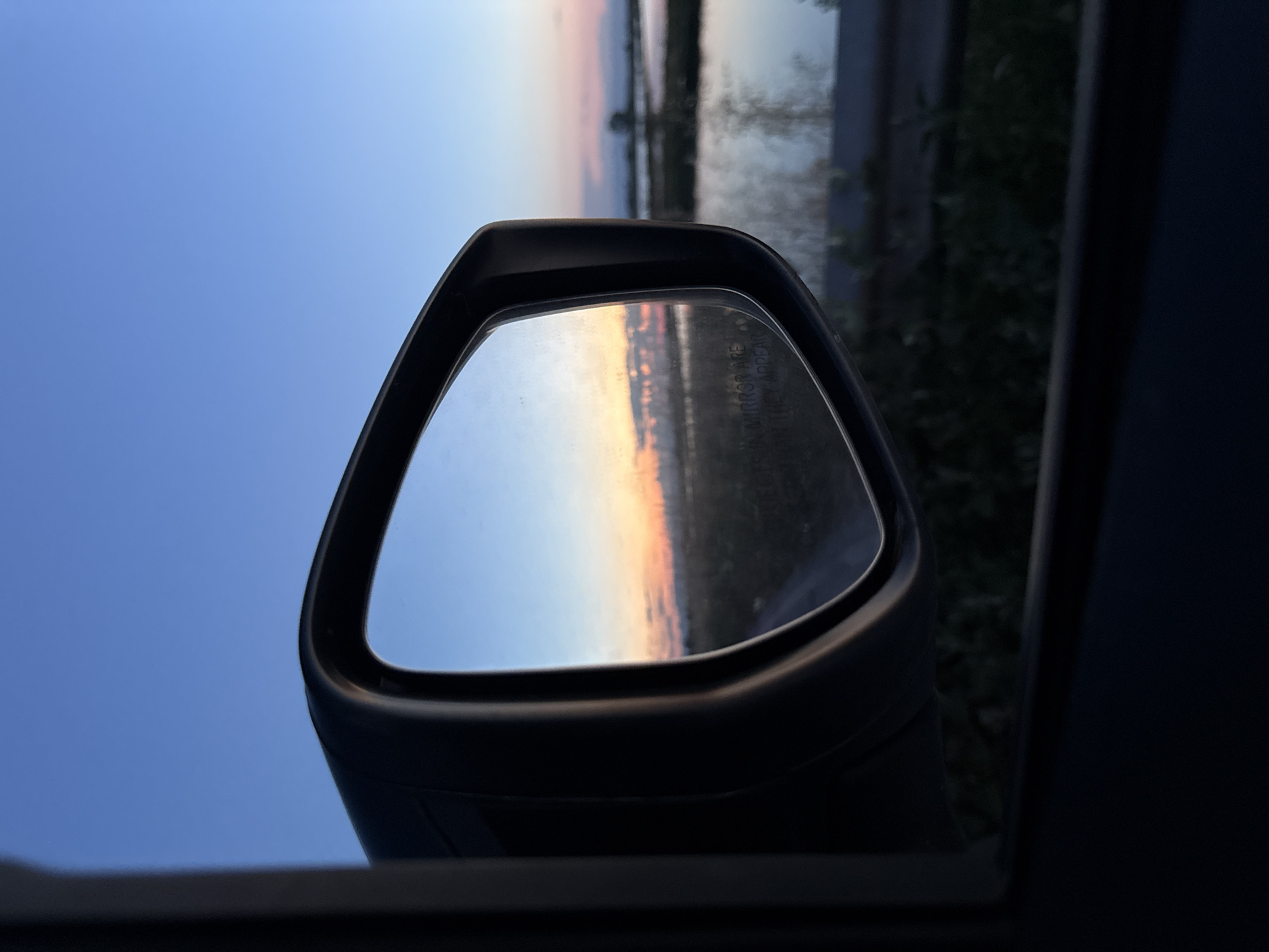 The image is a close-up shot of a car's side mirror reflecting a sunset over a body of water. The sky is a gradient of blue and orange, with soft clouds. The reflection shows a flat landscape with what appears to be a road leading into the distance. The water in the reflection is calm and reflects the colors of the sky. The car's interior frame is visible in the foreground, partially obscuring the view. The time of day is dusk or dawn. The location is not specified in the image, nor is the city or state of Unknown. There are no people or animals present in the image. The overall scene evokes a sense of peace and tranquility.