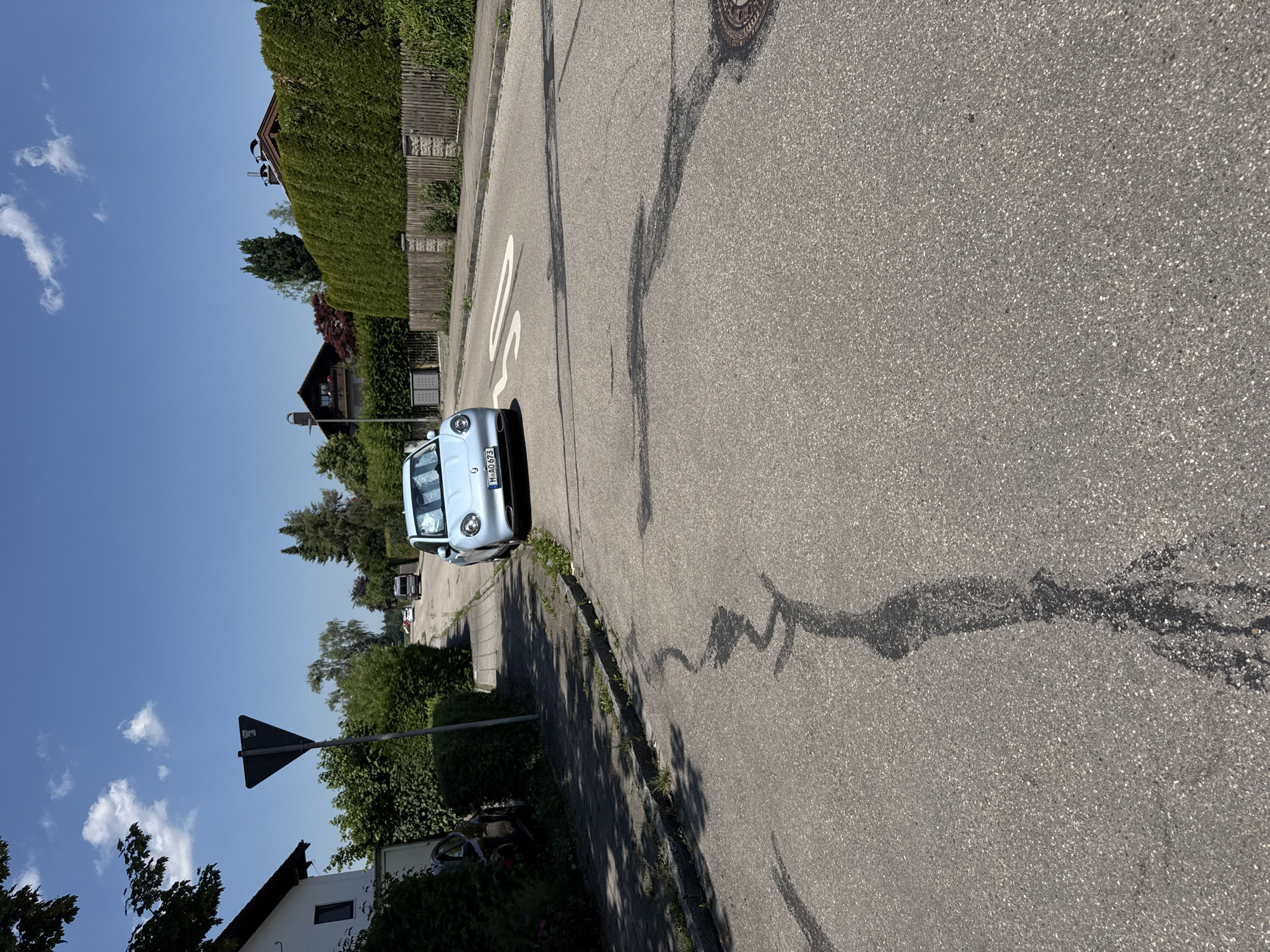 This media file shows a street scene in Höhenkirchen-Siegertsbrunn, Germany. The primary objects are a light blue car parked on the side of a paved road, a traffic sign, and residential buildings with green hedges and fences. The car has a license plate that reads "M-AO 673". The road has some cracks and dark patches, and a white line marking is visible. The setting is a residential area with trees and lush greenery. The sky is clear blue with a few white clouds, suggesting a pleasant day. The lighting indicates it is daytime. There are no people visible in the image. The overall impression is one of a quiet, suburban street.