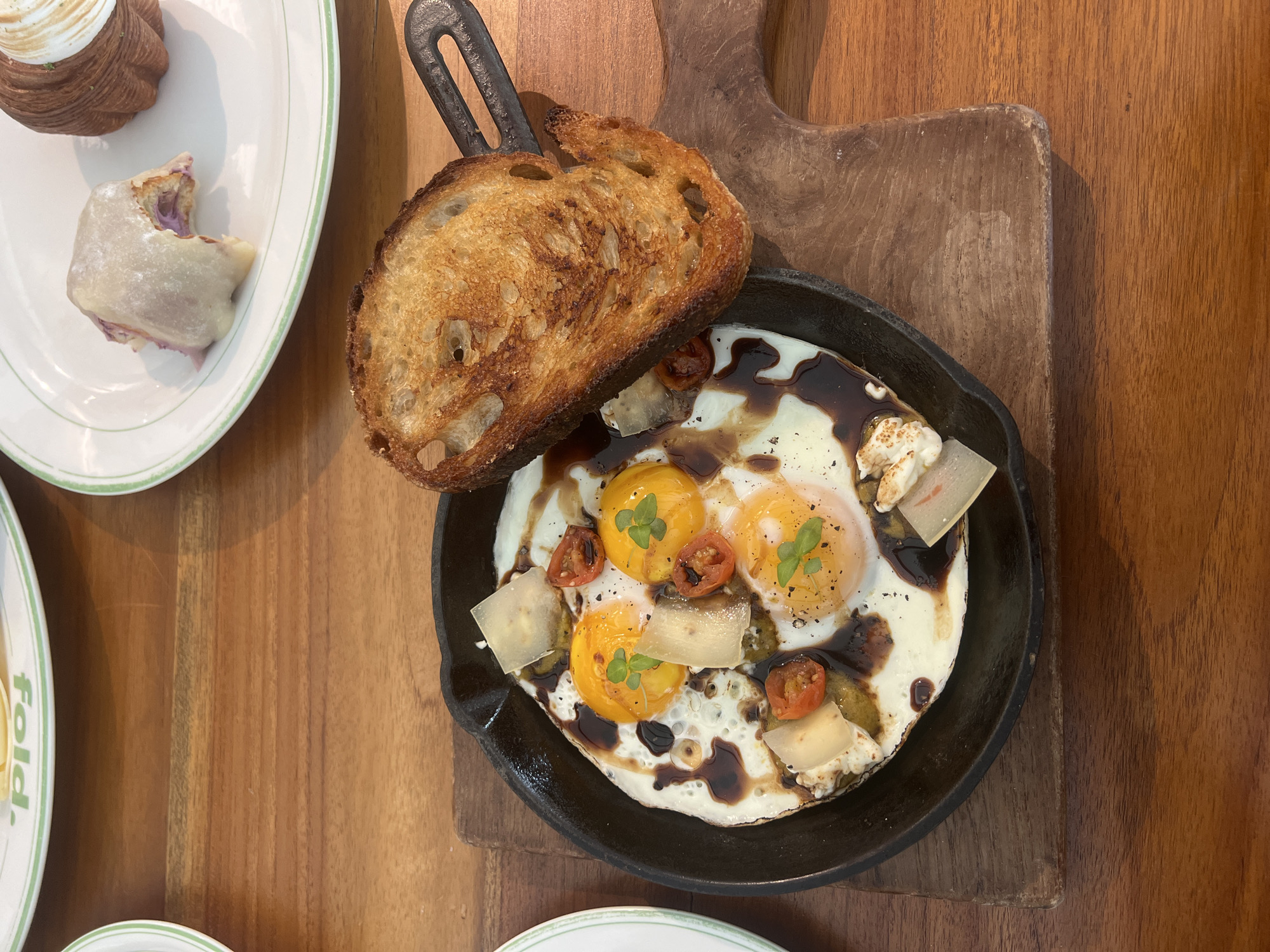 The image depicts a breakfast scene on a wooden table, likely in a restaurant setting. In the upper left, a white plate with a pastry and a dessert is visible. The main focus is a small cast iron pan containing four fried eggs, topped with cherry tomatoes, cheese slices, and basil, drizzled with a dark sauce. A slice of toasted bread rests on the edge of the pan. The pan sits on a wooden cutting board. Partially visible at the bottom left are the rims of additional plates with a green trim, and the word "fold." North Kuta, Indonesia, is not mentioned or visible in the image.