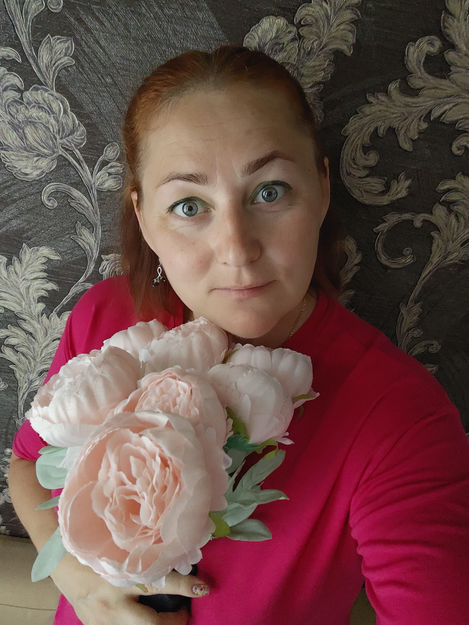 A woman with red hair and blue eyes is holding a bouquet of pink peonies in front of her. She is wearing a bright pink shirt. The background is wallpaper with a floral pattern. The scene is likely indoors, possibly a home, in Saint Petersburg, Russia.