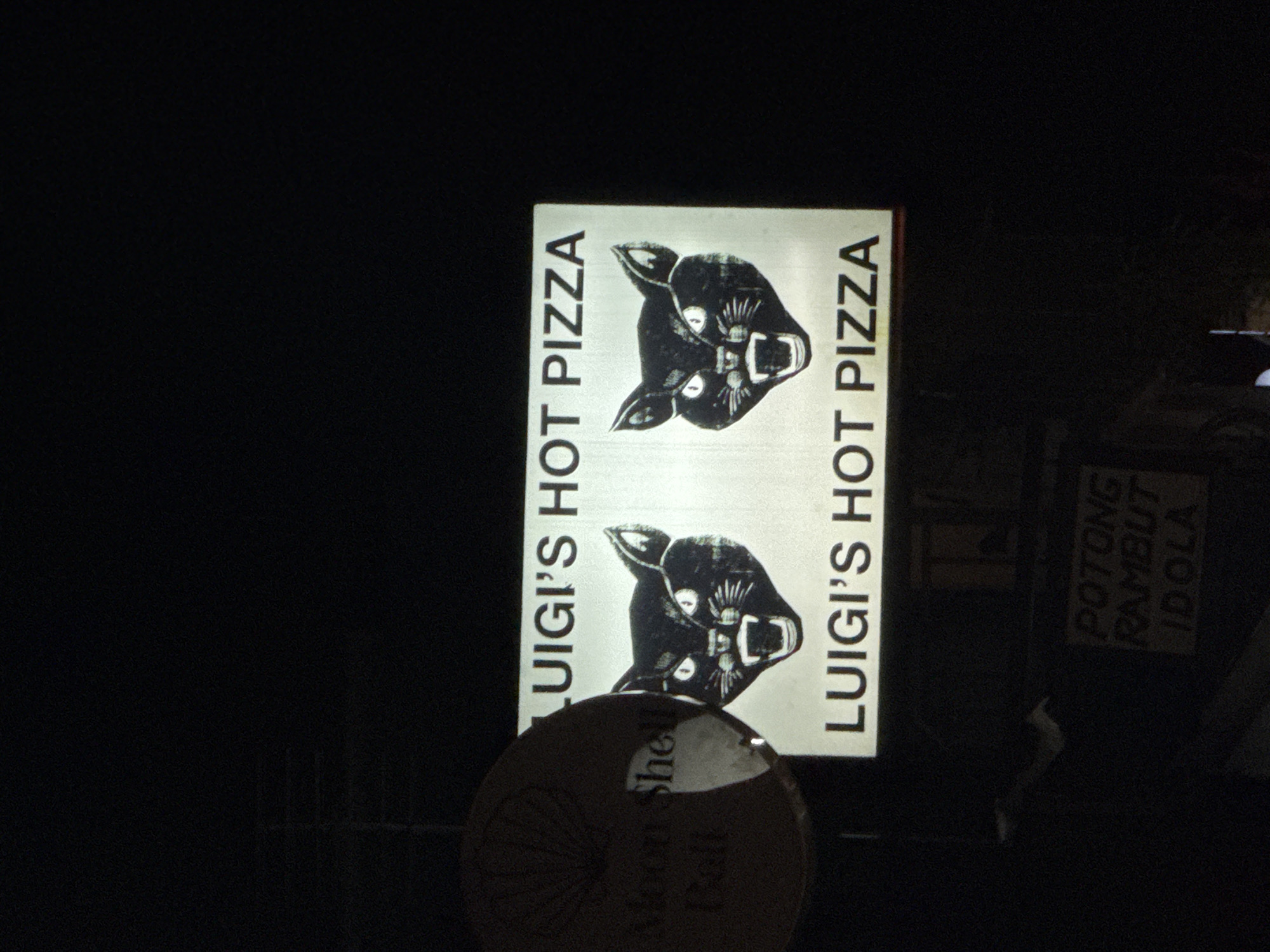 The image shows a sign for "Luigi's Hot Pizza" in North Kuta, Indonesia. The sign is white with black text and contains a graphic of a panther's head. Beneath the sign is a round object, likely a promotional item, with faint text partially visible. To the right of the pizza sign is another sign with the words "POTONG RAMBUT IDOLA." The background is mostly dark, suggesting it is nighttime. The perspective indicates the photo was taken at a low angle, looking up at the signs.