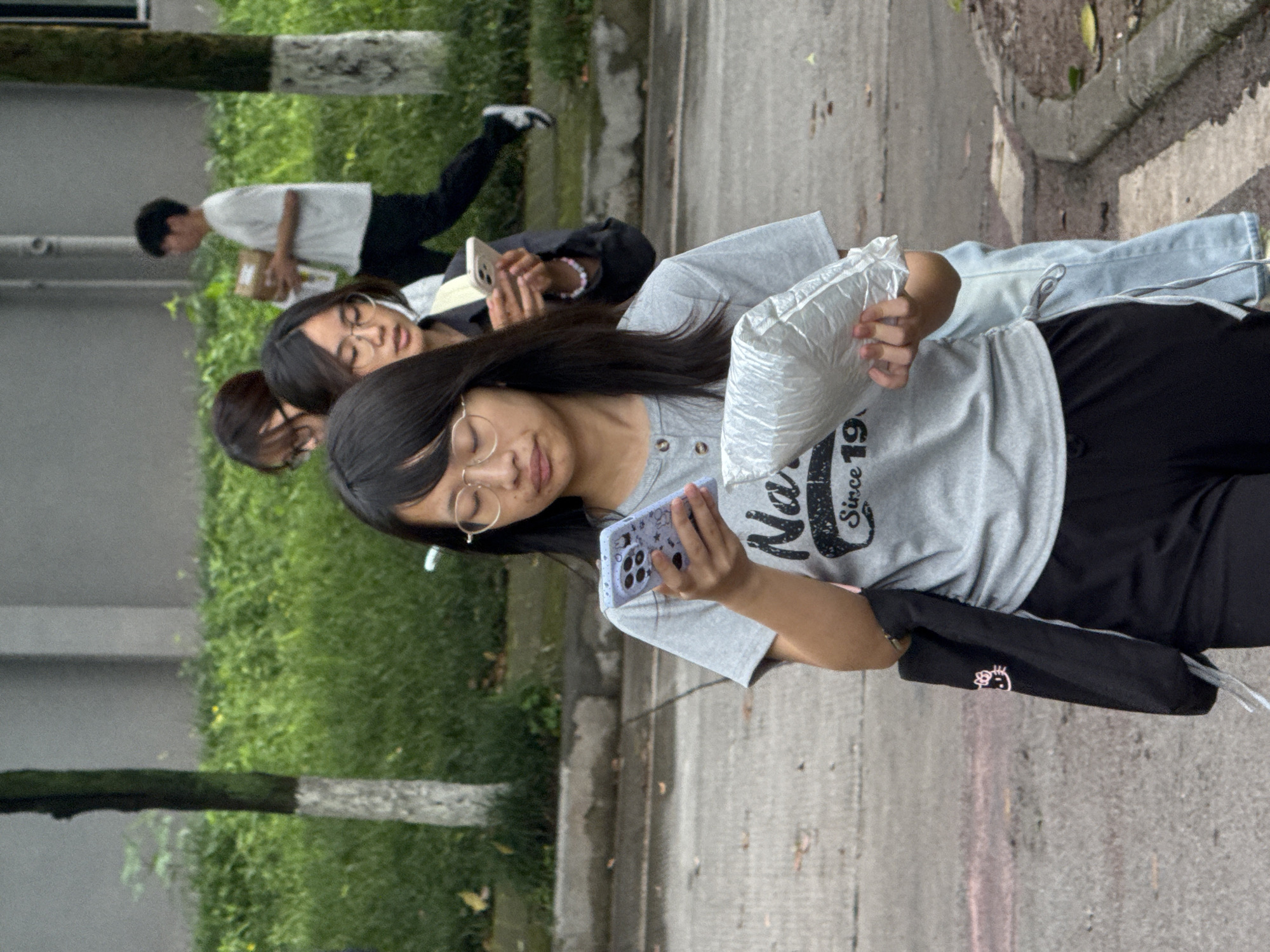 Captured in what appears to be an urban setting, the image displays a group of individuals in motion. A young woman, wearing glasses, is prominently featured; she is holding a smartphone in one hand and a crumpled plastic bag in the other. Two other young women are behind her, and a young man is in the distance. The backdrop shows trees and a paved path, suggesting a park or campus environment. The exact location within Jiangjin, China, cannot be determined from the image alone.