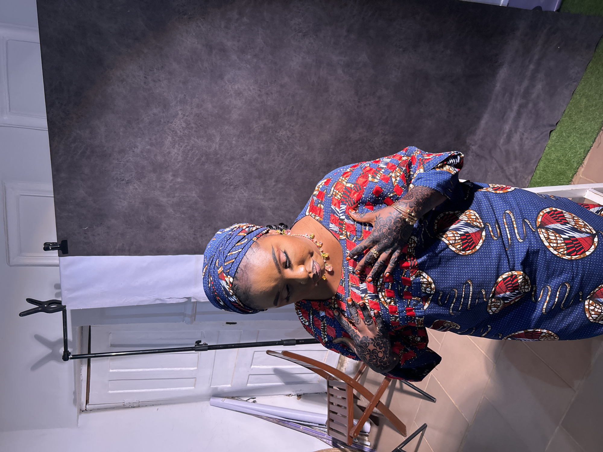 A medium indoor shot depicts a woman with medium skin tone and dark hair, wearing a navy blue, red, and white patterned garment and a blue head wrap, standing with her head and hands bent, possibly in a studio setting. Her hands, wrists, and upper chest are decorated with dark-colored henna. She also wears a necklace. Behind her, there's a grey backdrop, and to the left, there's a white wall with a photo studio setup that includes a stand and backdrop holder. A folding chair is nearby. The setting seems to be in a controlled environment, with artificial lighting being used. The location appears to be an indoor photo studio in Jos, Nigeria, based on the woman's dress and jewelry.