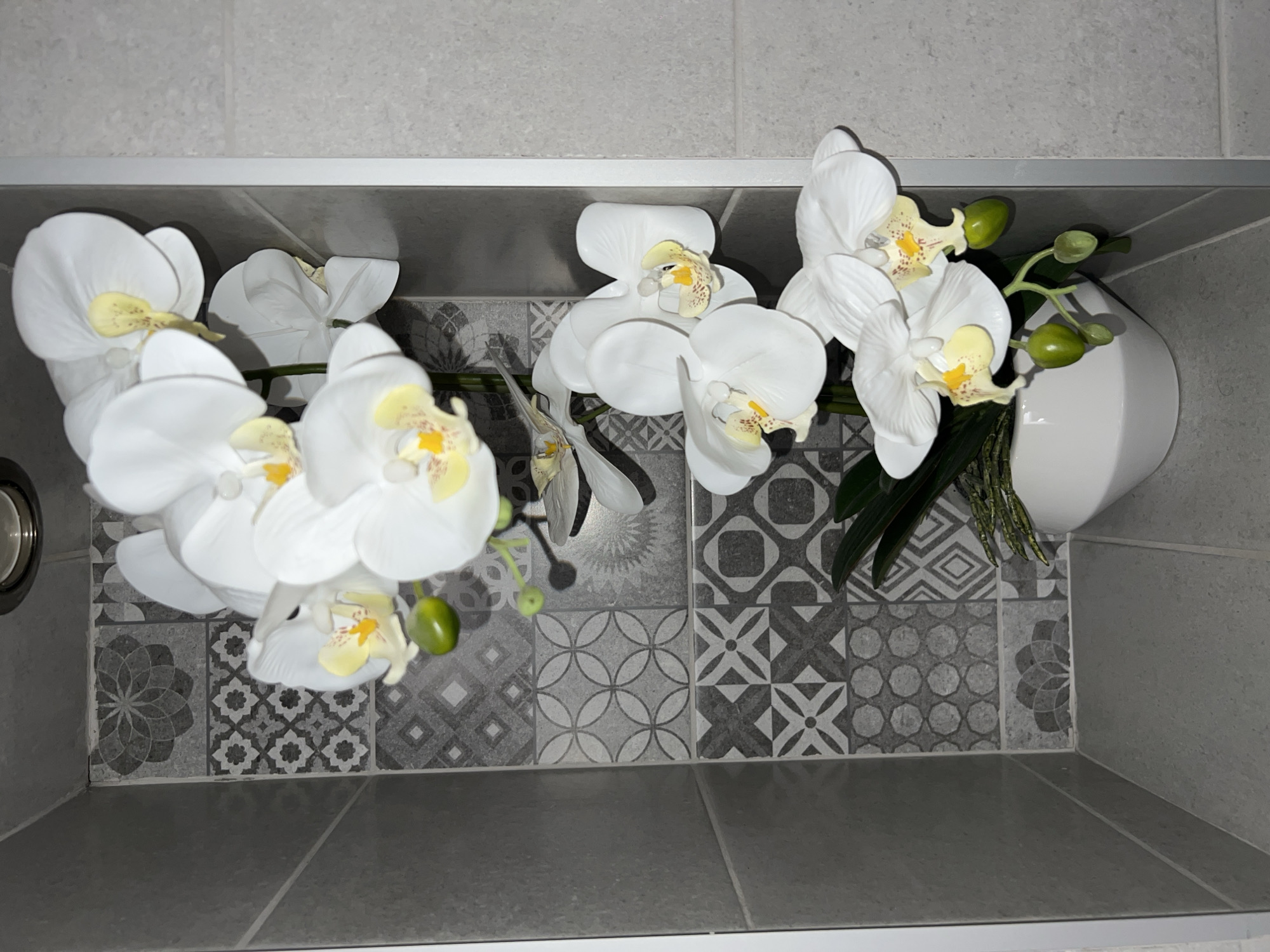 A still-life photograph shows a decorative arrangement of artificial white orchids in a white vase, placed in the corner of a bathroom in Burgas, Bulgaria. The background consists of grey, patterned wall tiles, with additional plain grey tiles above and below. The composition is an overhead shot.