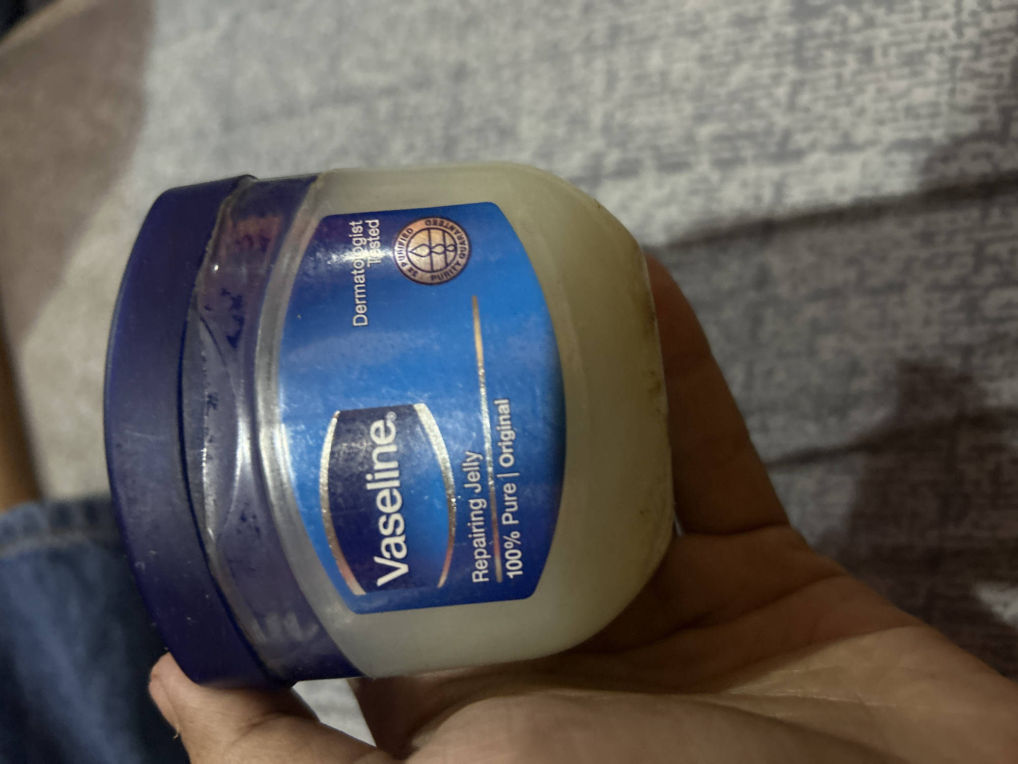 A hand holds a small, clear plastic jar of Vaseline petroleum jelly, with a blue lid. The label on the jar is primarily blue and white, displaying the Vaseline logo, "Vaseline", "Repairing Jelly", and "100% Pure | Original". There is also a circular emblem indicating "3x Purified" and "Purity Guaranteed". The background appears to be a textured gray surface, possibly a blanket or fabric. The lighting suggests an indoor environment. There is no indication of the city of Depok, Indonesia in the image.