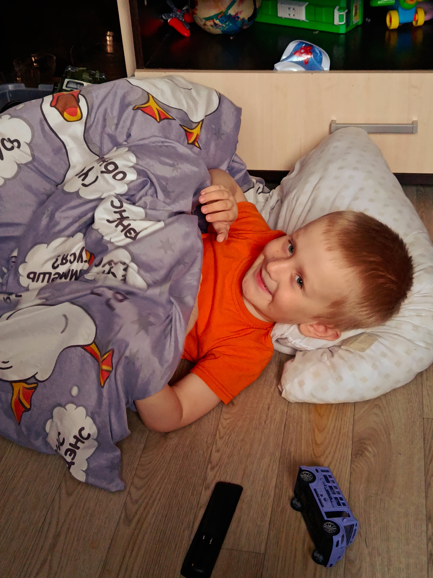 An indoor scene captures a young child, likely a boy with light brown hair and blue eyes, lying on a light wooden floor. He is wearing a bright orange t-shirt and is covered up to his waist by a purple blanket decorated with white cartoon geese, yellow stars, and scattered Cyrillic text. His head rests on a white, subtly patterned pillow, and he is looking upwards and smiling widely towards the camera, conveying happiness and playfulness.
On the wooden floor nearby, a black remote control is visible, along with an overturned purple toy car resembling a minivan or SUV. In the background, a light-colored wooden cabinet or dresser stands, featuring a silver pull handle. Above this, a dark shelf holds various items, including a green plastic toy container, a small red and blue object, and a multi-colored spherical item, possibly a globe or ball.
The setting appears to be a child's bedroom or play area. The Cyrillic text on the blanket includes discernible fragments such as "ОНЧ" and "УГО," along with other partially visible or inverted letters like "СВЯ" and "ЯЩА," all presented within white cloud-like shapes. The overall atmosphere is cheerful and relaxed.