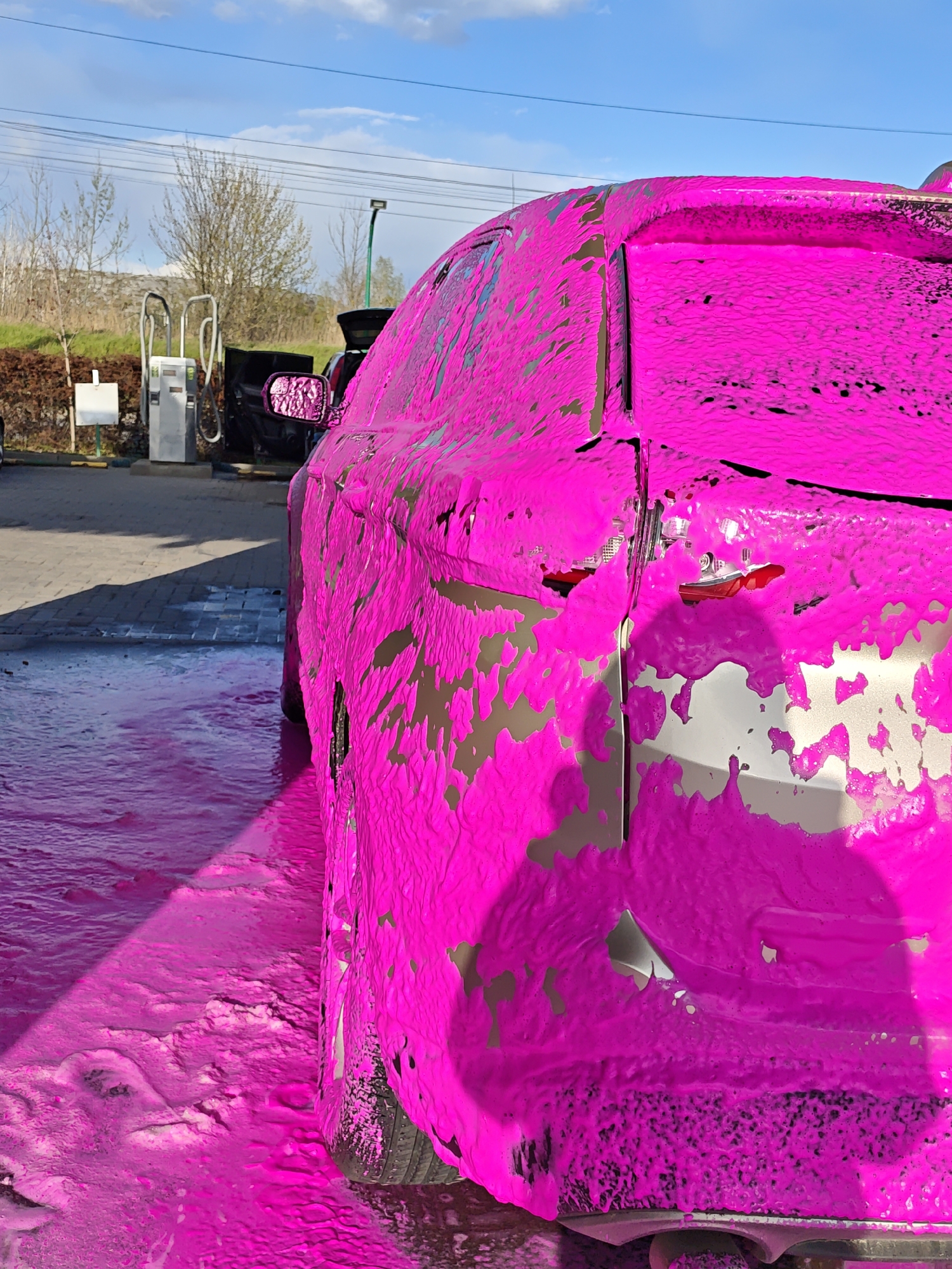 The image shows the rear of a silver car covered in bright pink foam, likely at a car wash. The location appears to be outdoors, with a clear, blue sky and some clouds. In the background, there are car wash service machines and some greenery suggesting a roadside location. The foam covers the entire visible part of the car, obscuring details. The foam is thick enough to be flowing on the ground and is visibly pooling in puddles. The area around the car appears to be paved. The location is in the city of Vinnytsia, Ukraine.