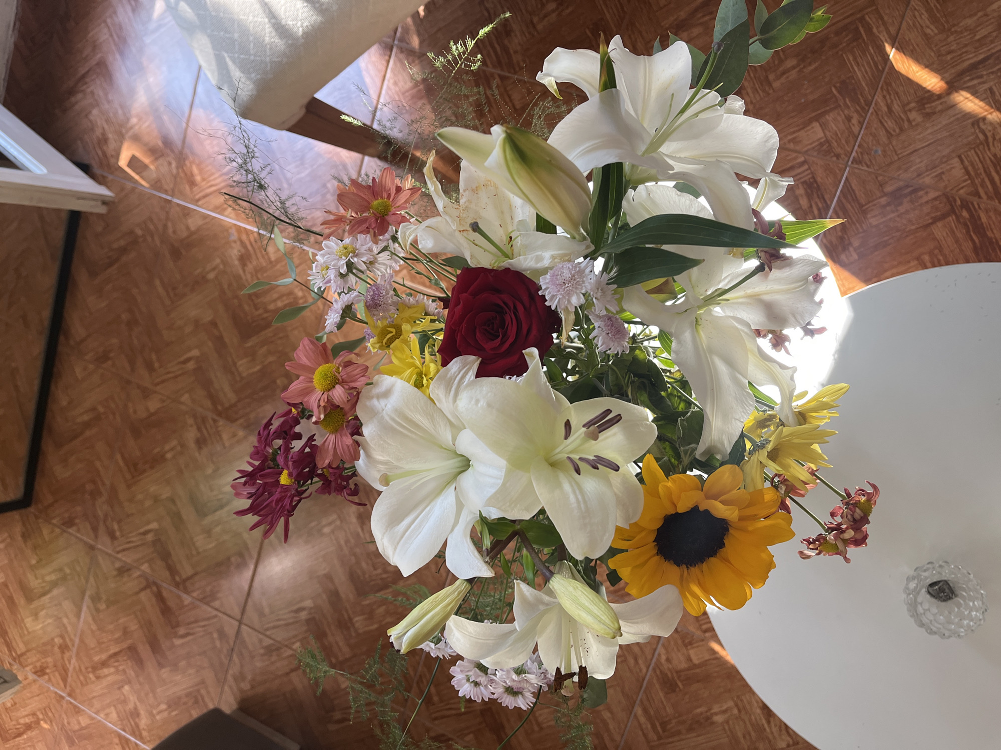 A top-down shot shows a bouquet of various flowers including white lilies, a dark red rose, sunflowers, and other colorful daisies, likely arranged in a vase or container not visible. The bouquet is positioned on a white, round table, and the surrounding environment includes a tiled floor and what appears to be a portion of a chair or piece of furniture. The lighting is bright and sunny, suggesting an indoor setting with natural light coming in from a window. There are no people visible in the frame, and the primary focus is on the floral arrangement. The location cannot be determined as San Fernando del Valle de Catamarca, Argentina, or any other specific location based solely on the visual information provided.
