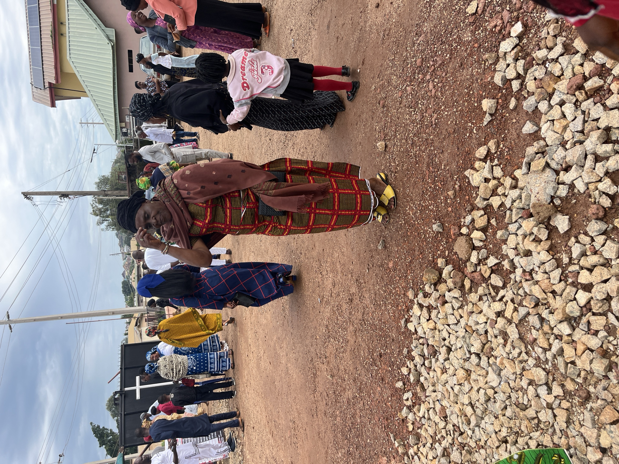 The image depicts a gathering of people outdoors, possibly a community event or a religious service, in Jos, Nigeria. The ground is covered in reddish-brown earth and loose gravel. In the background, several buildings are visible, including one with solar panels on its roof and another with a prominent white cross on a dark structure, suggesting a religious affiliation.
Several individuals are present, dressed in a variety of traditional African attire and modern clothing. A woman in the foreground is wearing a vibrant, checkered dress with a patterned shawl. To her right, a young girl is dressed in a pink sweatshirt with "Dreamer Girls" and the number "3" on it, a black skirt, and red tights. Other people in the image are seen in groups, engaged in conversation or observing their surroundings. The sky is overcast, indicating a cloudy day. The overall scene suggests a lively community event taking place in an urban or semi-urban setting.