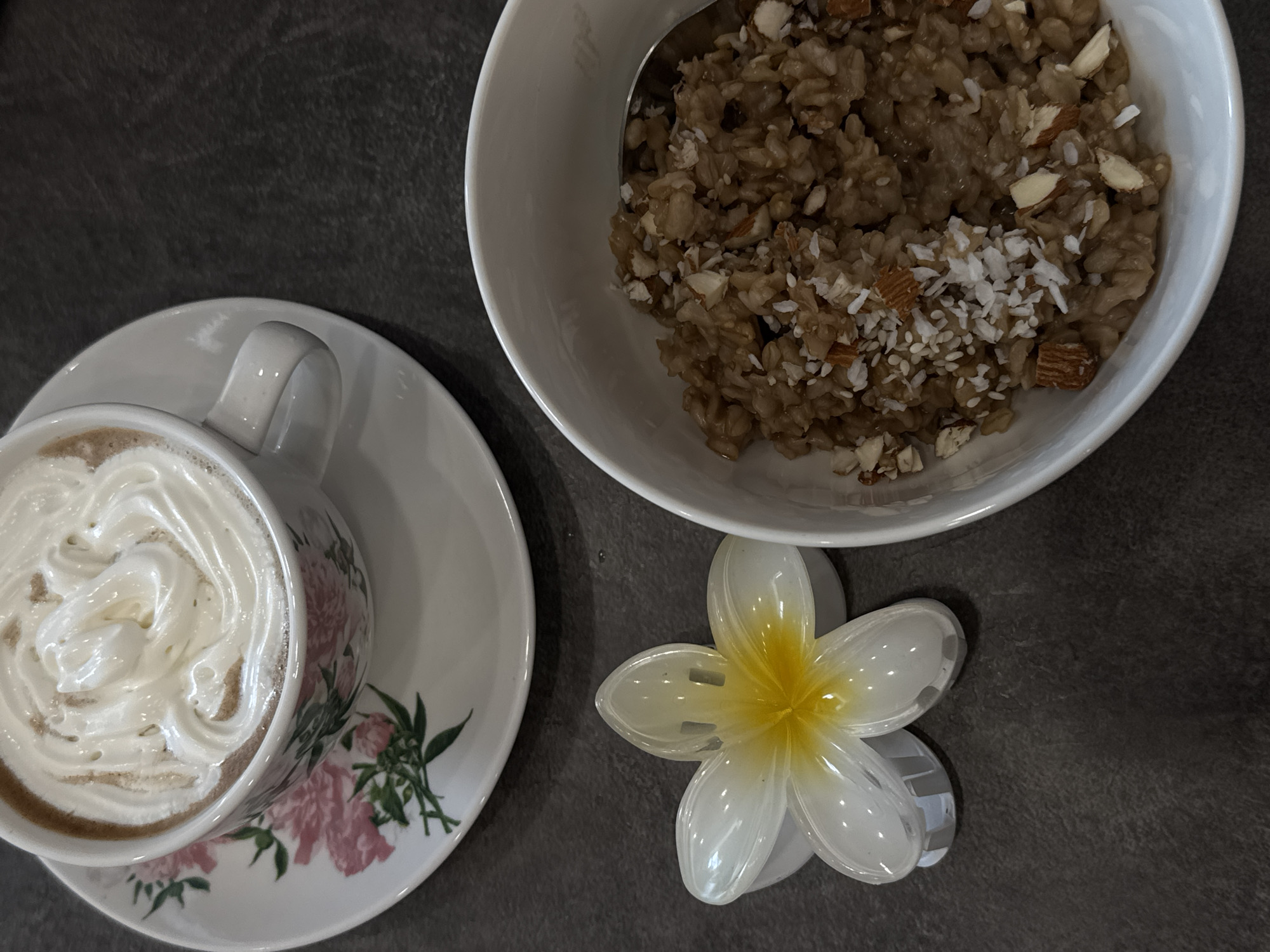 A close-up overhead shot shows a breakfast scene on a dark gray textured surface. In the upper left, a white teacup with a floral pattern of pink roses and green leaves sits on a matching saucer. The teacup is filled with a brown liquid, topped with a generous swirl of white whipped cream. To the lower left of the teacup, a white and yellow plastic flower-shaped hair clip is placed on the surface. To the right of the hair clip, a white bowl contains a portion of oatmeal. The oatmeal is a light brown color and is topped with chopped almonds and shredded coconut. A silver spoon is partially submerged in the oatmeal.