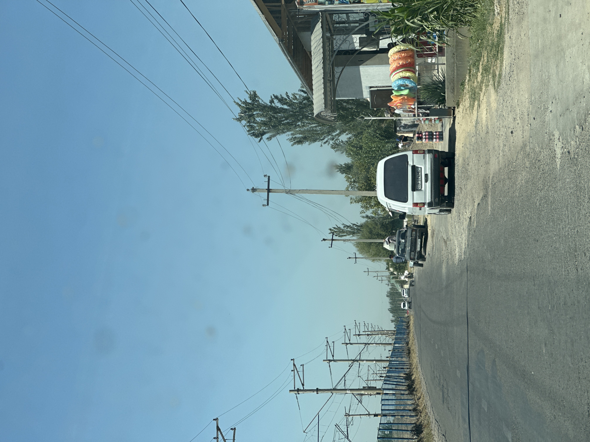 This image depicts a street scene in Erkin, Uzbekistan. The road is paved but appears worn in places, with visible tire tracks. To the left of the road, there is a blue metal fence bordering a grassy area, and beyond that, a row of utility poles with overhead wires stretches into the distance. The right side of the road features a building with a gray awning and an open doorway. In front of the building, a rack displays colorful inflatable rings and other merchandise. A white minivan is parked on the right side of the road, facing away from the camera, with its license plate visible. Further down the road, there are other vehicles, including a dark-colored car with a trailer. Several people are present, with some standing near the building and others on the roadside. Trees and utility poles are scattered throughout the scene, under a clear blue sky.