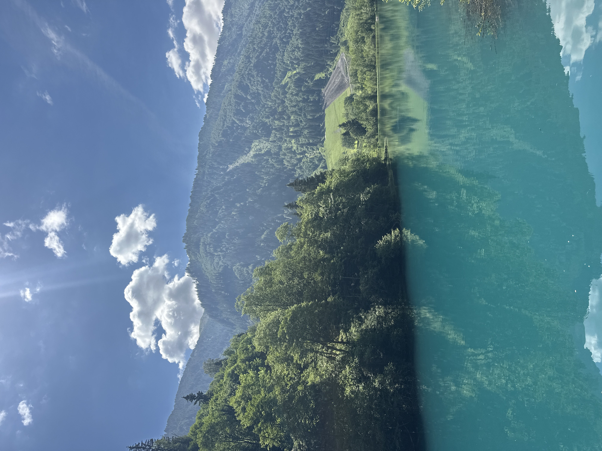 This is a vertical, full shot of the turquoise waters of the Klammsee lake in Kaprun, Austria. The lake dominates the foreground, with its still surface reflecting the lush green trees on the left bank and the verdant, forested mountains in the background. The sun is shining brightly, casting dappled light on the trees and creating sunbeams in the clear blue sky dotted with fluffy white clouds. A large section of the mountain on the right appears to have been cleared for construction or development, revealing a lighter green patch against the dense forest. The scene is serene and picturesque, characteristic of the Austrian Alps.