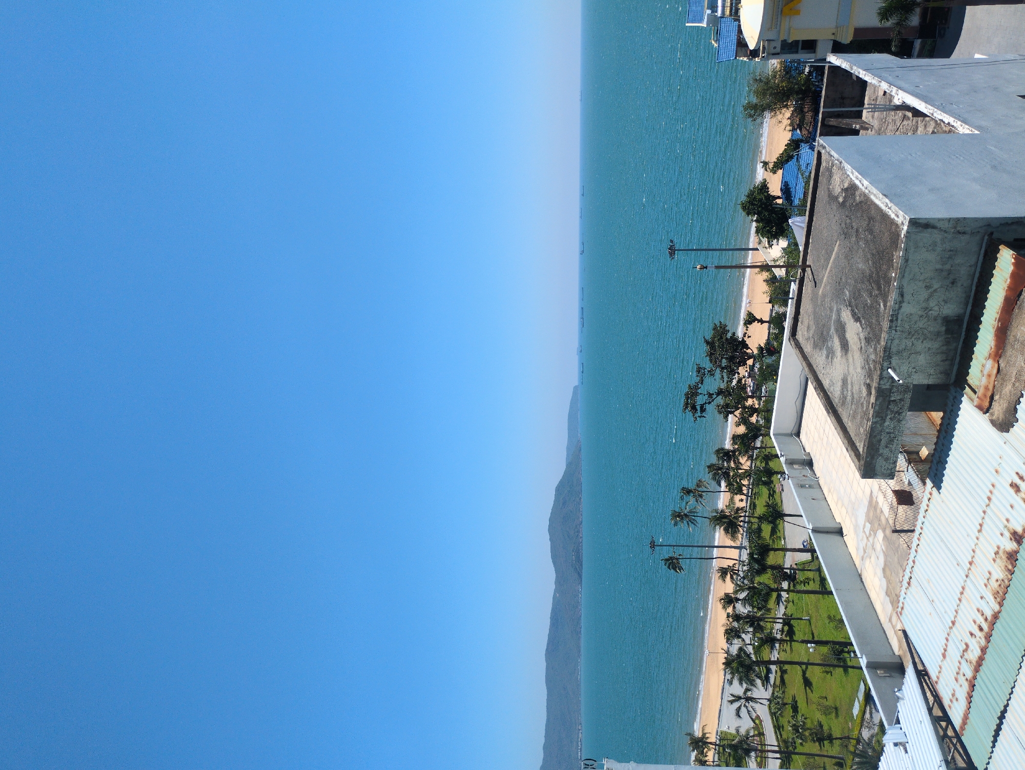 The image depicts an elevated view of a coastal scene in South Quy Nhon Ward, Vietnam, under a clear, bright blue sky. In the immediate foreground, multiple rooftops are visible, featuring weathered corrugated metal and flat concrete surfaces with perimeter walls. One flat concrete roof exhibits dark stains and dirt.
Beyond the rooftops, a landscaped beachfront area runs parallel to the coastline. This area includes a paved road, a strip of green grass, and numerous mature palm trees. Several tall streetlights are positioned along this strip. A light-colored sandy beach separates this parkland from the ocean.
The midground is dominated by a vast expanse of turquoise-blue sea, exhibiting a choppy surface with visible waves. Along the distant horizon of the ocean, approximately a dozen large cargo vessels or tankers are discernible as dark silhouettes. In the far background, a range of verdant mountains extends across the left side of the horizon.
No human figures are visible on the beach, in the park area, or on the rooftops. The scene appears illuminated by bright sunlight, indicative of daytime.