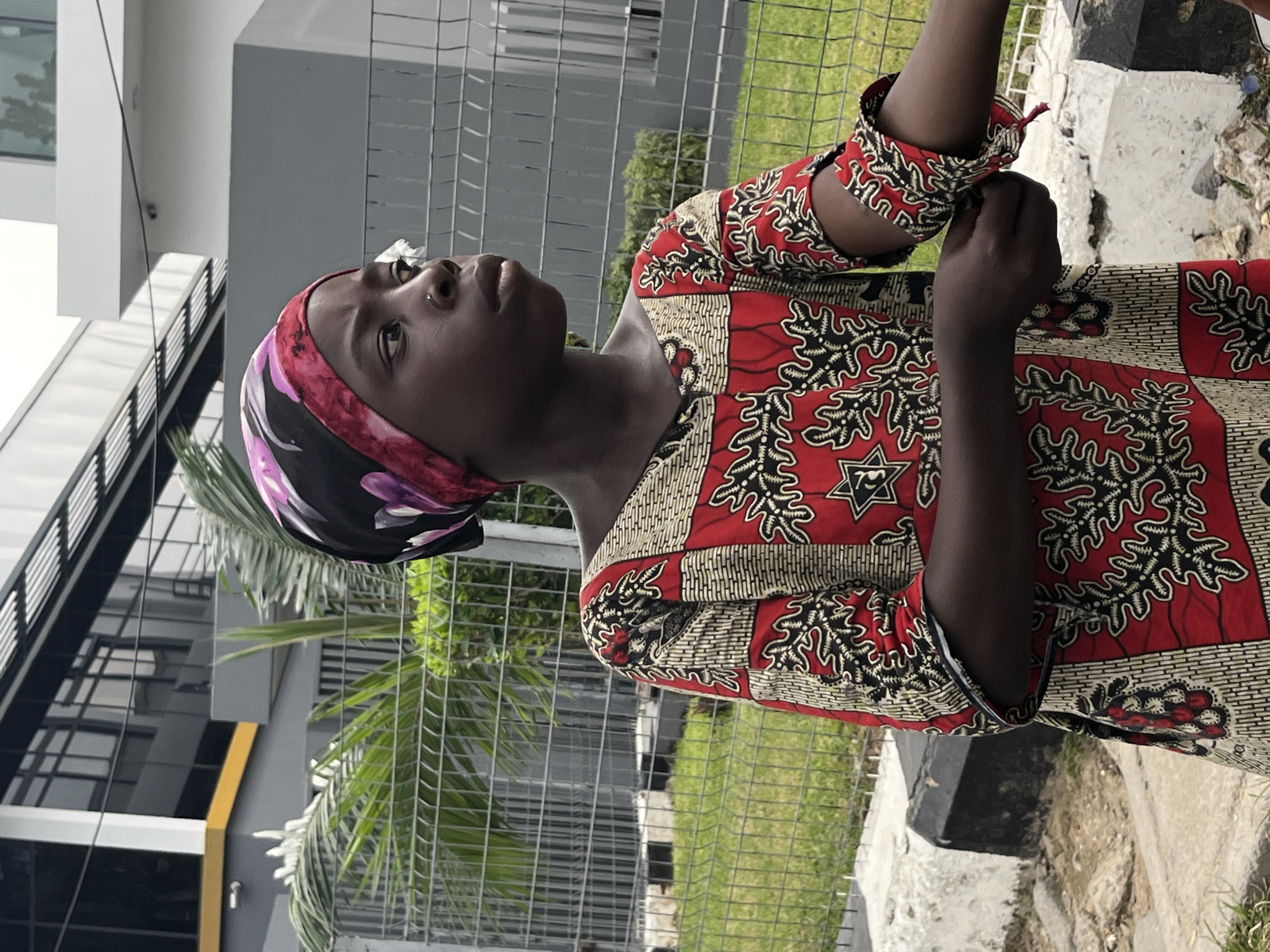 A young woman in a red, black, and gold patterned dress and a floral headscarf stands outside in Itirin, Nigeria. She is looking upward. She has tied off the wrist cuff of her right hand on the dress. She stands in front of a chain-link fence and behind a concrete barrier. A modern gray building is visible through the fence. Green grass and the fronds of palm-like plants are visible behind the fence.