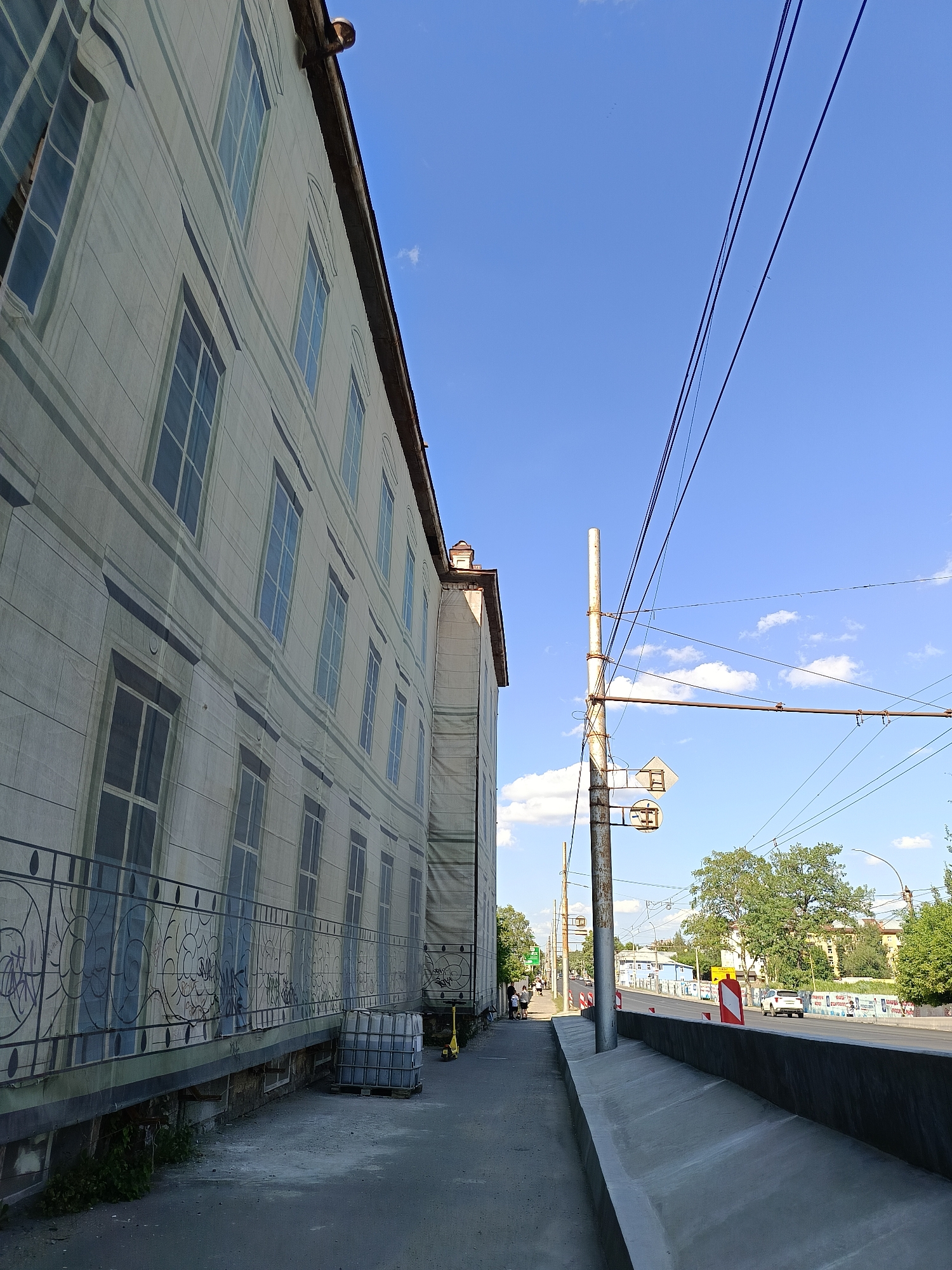 The image shows a street scene with a building on the left and a road on the right. The building is covered with a large banner that mimics the appearance of a historical facade with numerous windows. A sidewalk runs alongside the building, bordered by a decorative metal fence with graffiti. On the sidewalk, a large blue plastic container on a pallet is visible, along with a yellow scooter. A street light pole with traffic lights and overhead wires stands on the right side of the sidewalk. The road is visible in the background, with cars and trees lining its sides. The sky is clear blue with a few scattered clouds. The scene suggests an urban environment, possibly undergoing construction or renovation, given the banner and the construction materials.