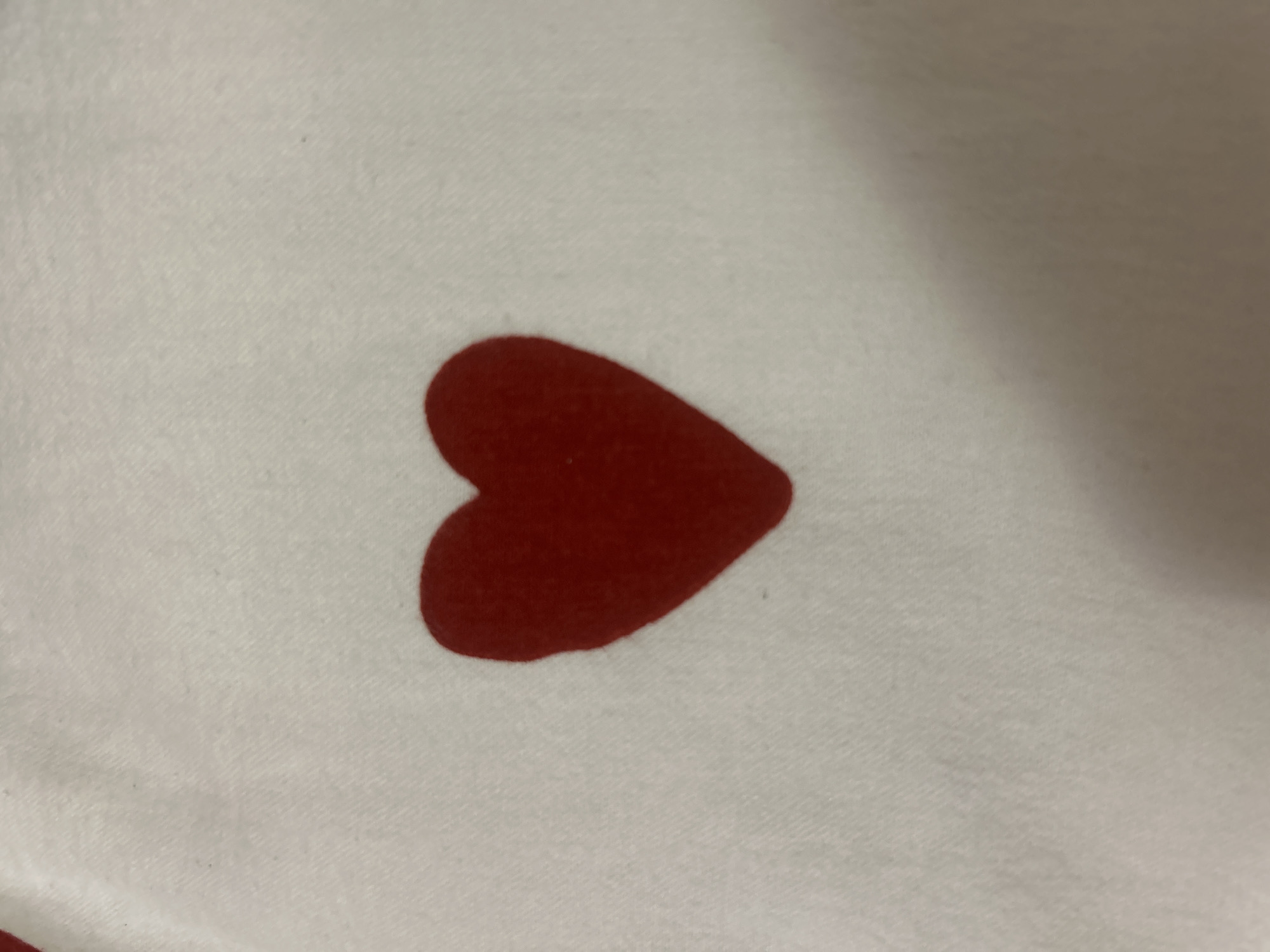 The image shows a close-up view of a red heart shape on a white fabric. The heart is solid red and appears to be a print or appliqué on the material. The fabric has a textured appearance with visible threads. The lighting is somewhat dim, creating soft shadows and highlighting the texture of the fabric. There are no people or other objects visible in the frame. The location in Krakow, Poland, is not discernible from the image itself.
