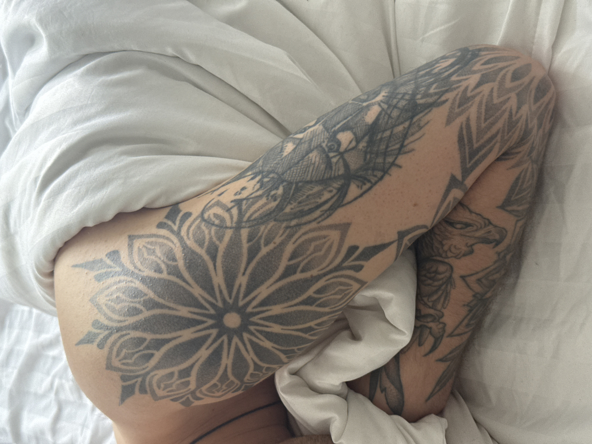 A pair of tattooed legs lies on a white sheet. The tattoos feature geometric patterns, a lotus flower, and a griffin. The scene is indoors. The location is not identifiable within the image.