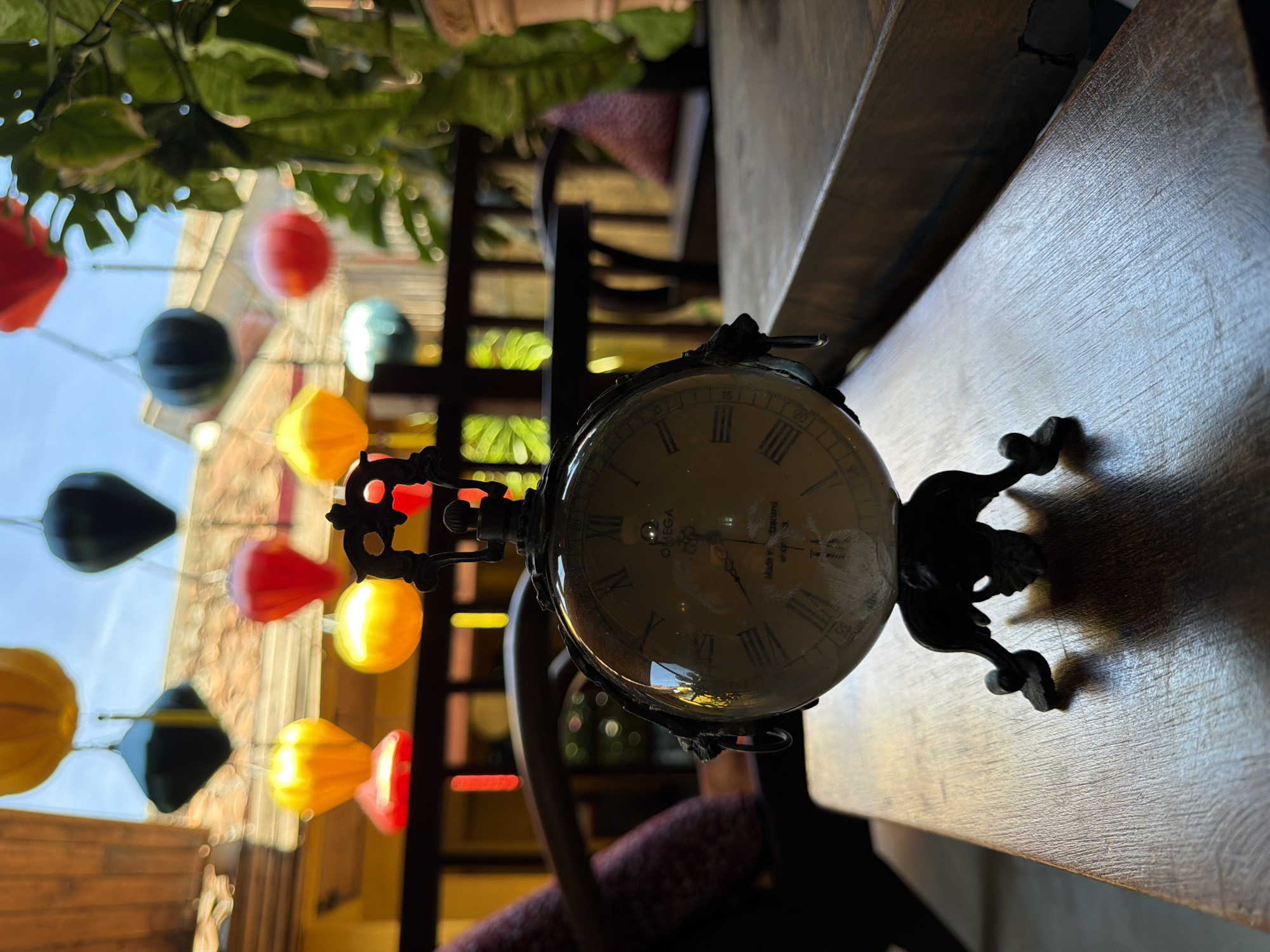 An antique-style pocket watch rests on a wooden table. The watch has a Roman numeral dial and an ornate metal stand. In the background, colorful paper lanterns hang from wires, creating a festive atmosphere. The scene is set in Hội An Ward, Vietnam, a city known for its historic architecture and vibrant street life. The lighting suggests it is daytime, with sunlight filtering through the lanterns and casting shadows on the table.