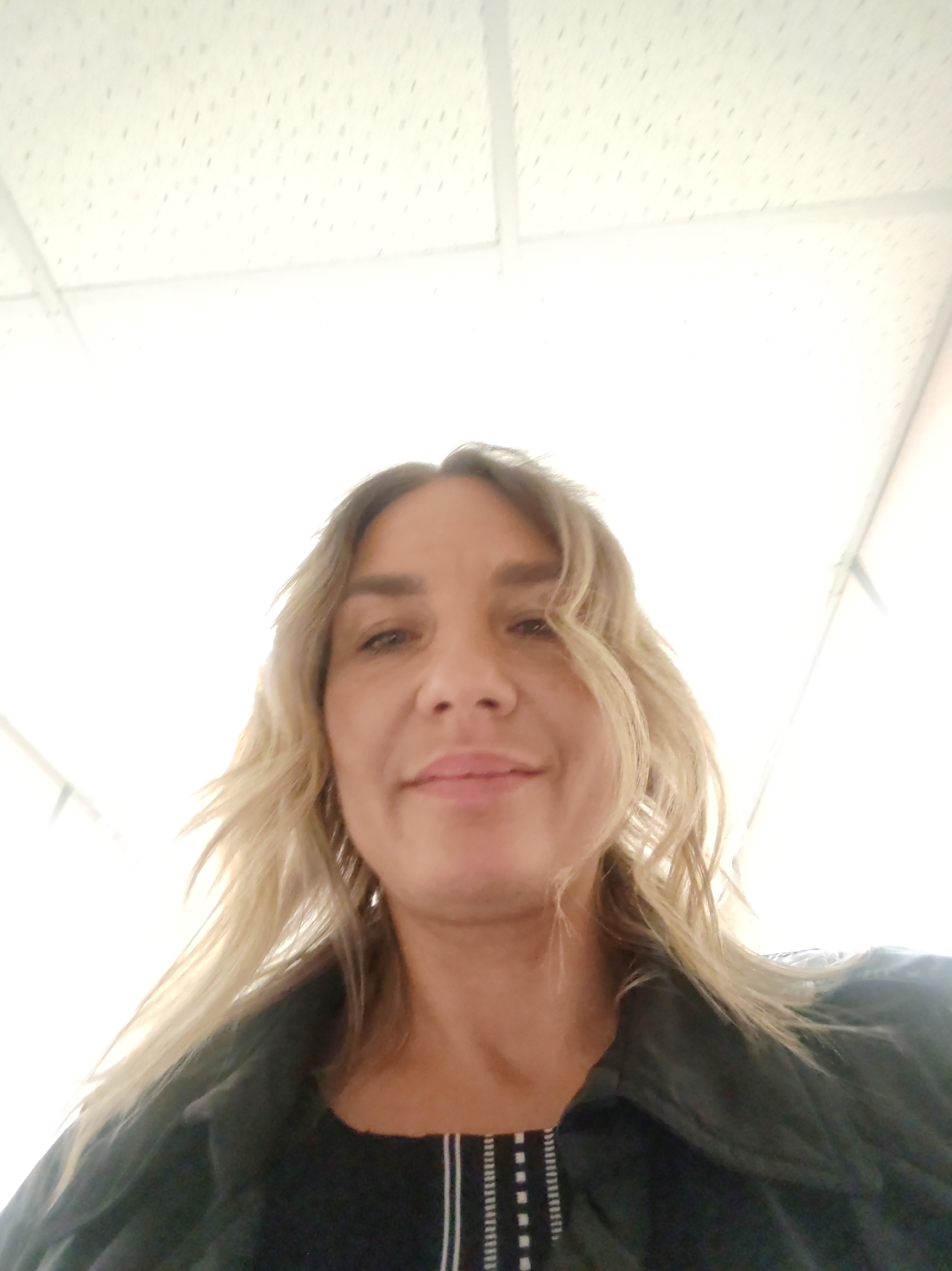 A woman with blonde hair and a black jacket is positioned in front of a white ceiling with a grid pattern. The location appears to be indoors in a room with white walls. The city is unknown.