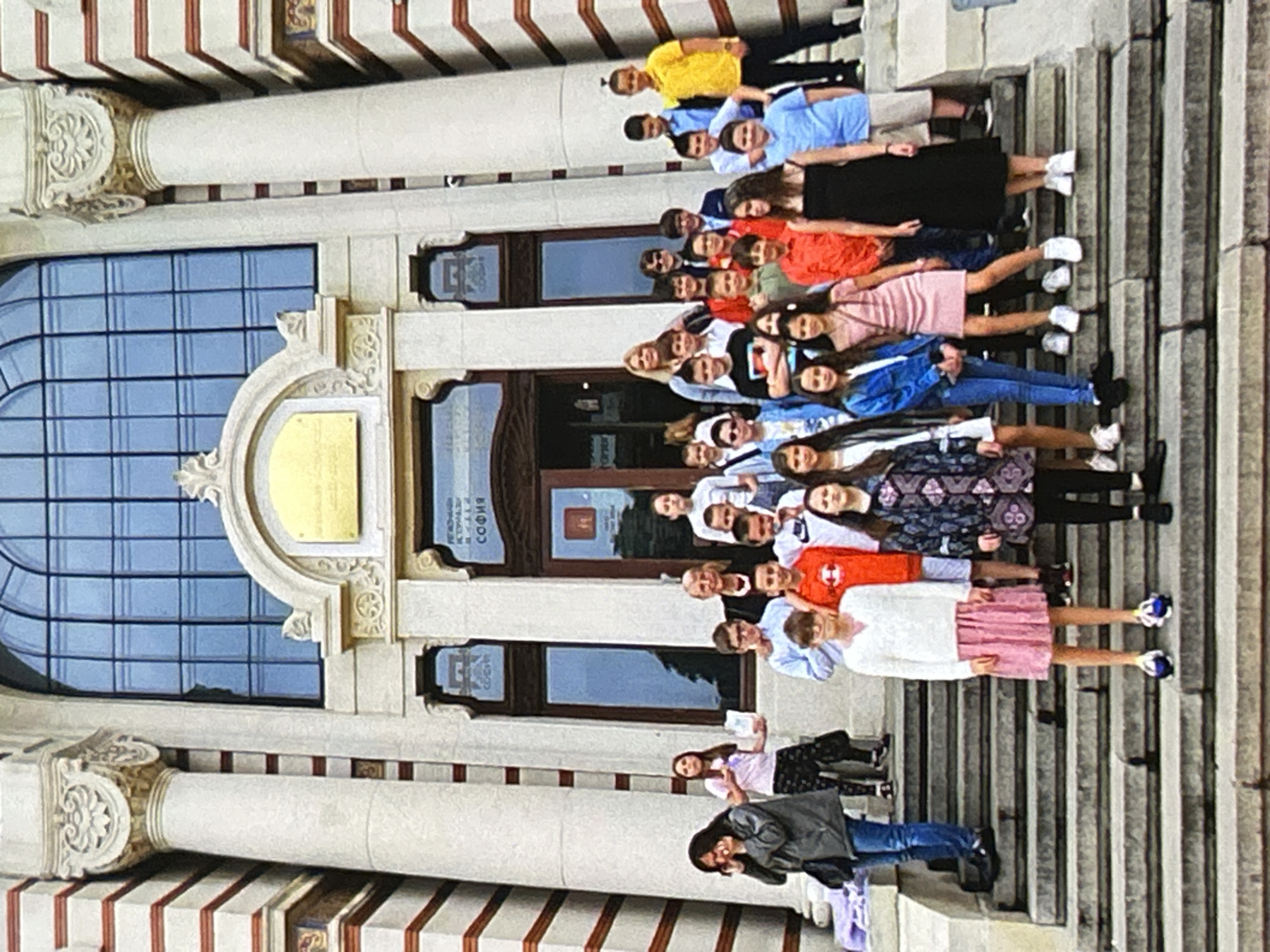 A group of approximately 30 individuals, predominantly children and teenagers, with a few adults, are assembled on the wide stone steps leading up to the entrance of the National Library "St. St. Cyril and Methodius" in Sofia, Bulgaria. The building's facade is characterized by light-colored stone, large classical columns with ornate capitals, and horizontal red-brown stripes. A prominent arched window with dark grid-patterned panes is visible above the central entrance, which features two wooden doors with glass panels. Text in Cyrillic, including "СОФИЯ," is visible on signs above and on the doors.
In the foreground, standing on the lowest visible step, are two individuals: a female wearing a white long-sleeved top and a pink pleated skirt with dark and blue athletic shoes, and to her right, a female in a dark blue and patterned jacket over a dark top and black leggings, also wearing dark shoes. Behind them, on higher steps, are numerous children and teenagers, mostly facing the camera, suggesting a group photograph. Their attire varies from casual t-shirts and jeans to dresses and collared shirts in various colors.
Several adults are interspersed among the younger group, particularly in the middle and upper sections. On the far left, an adult female with dark hair is standing on a lower step, holding a phone to her ear. On the right side, a tall male in a yellow polo shirt and black pants stands on a higher step, looking towards the camera. Another adult female with blonde hair is visible in the upper-middle section, wearing a light blue top and dark blazer. The scene is illuminated by daylight, indicating an outdoor setting. The individuals are positioned across the breadth of the steps, with a few small purple items visible on the far left of the lowest step.