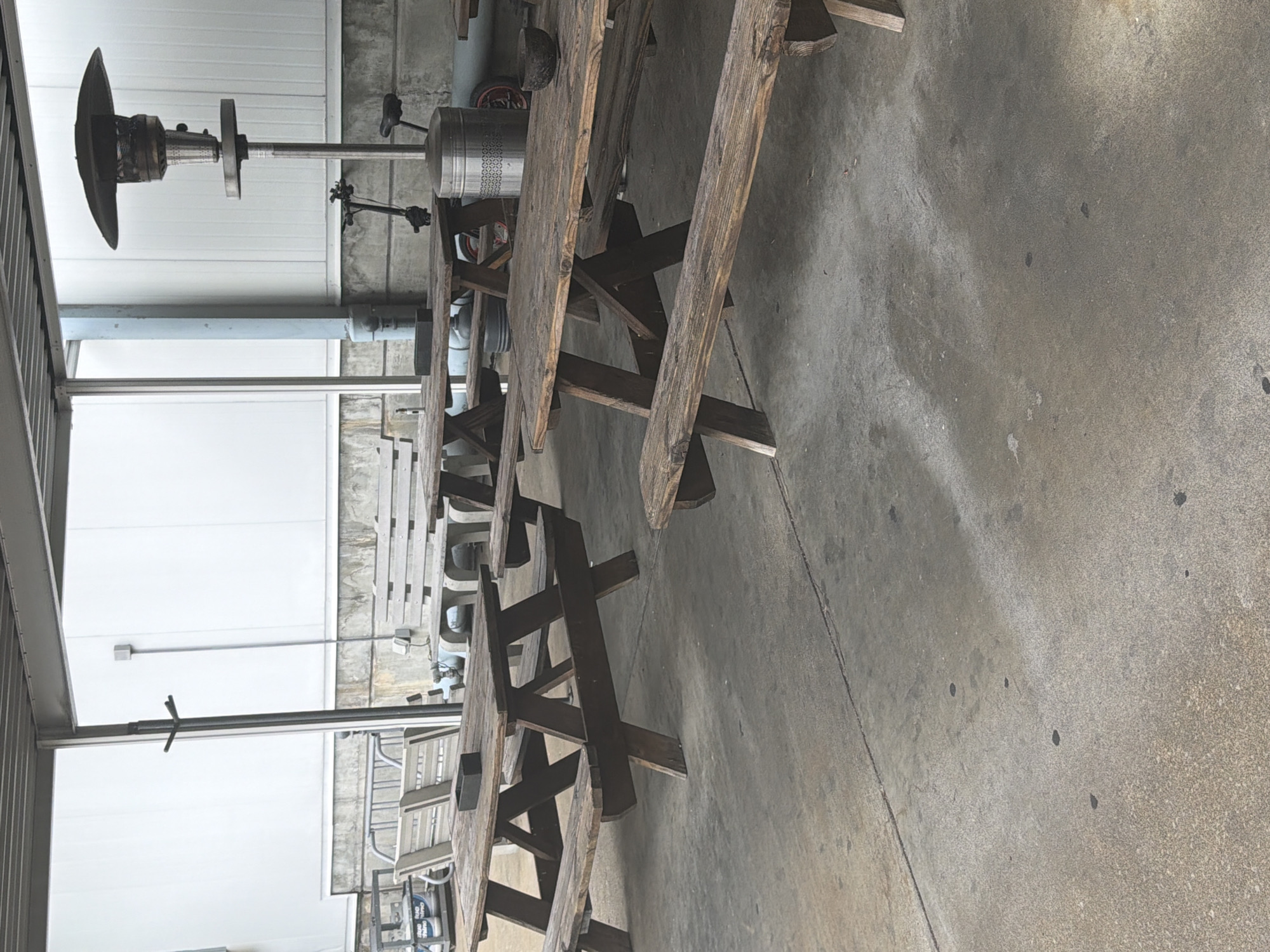 An outdoor seating area with several wooden picnic tables and benches is shown. A patio heater stands in the background, along with a small scooter. The ground is concrete, and the area is covered by a corrugated metal roof supported by metal poles.