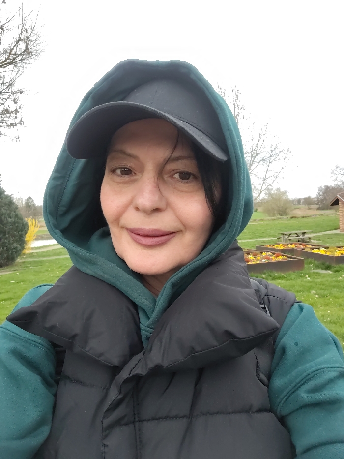 An adult female is centrally positioned in a close-up outdoor photograph, looking directly at the camera. She wears a black baseball cap, a dark teal hooded sweatshirt with the hood up, and a black puffer vest. Her dark hair is visible beneath the cap and hood, and her lips are colored a muted reddish-pink. The background features a green grassy expanse. To the right, multiple rectangular raised garden beds filled with yellow, orange, and red flowers are visible, along with a wooden picnic table with benches. Further back, a small building with a brown roof, bare trees, and distant vegetation are present under an overcast sky. The scene is located in a park or garden area within Souffelweyersheim, France.