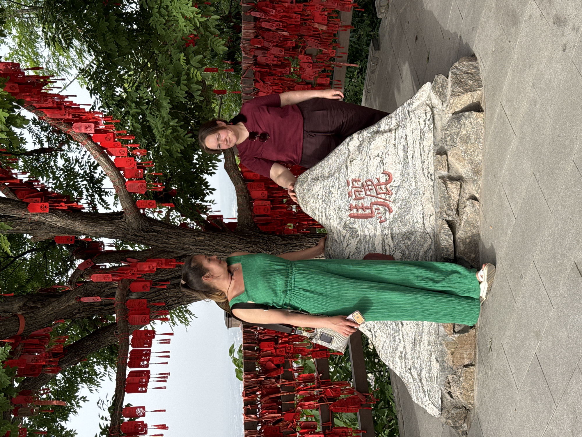 Two women pose near a rock with Chinese writing under a tree adorned with red tags in the Lintong District of China. One woman is dressed in a green jumpsuit and holds a phone. The other wears a burgundy top and dark pants, both touching the rock. The background features lush greenery, a paved area, and a hint of a distant view.