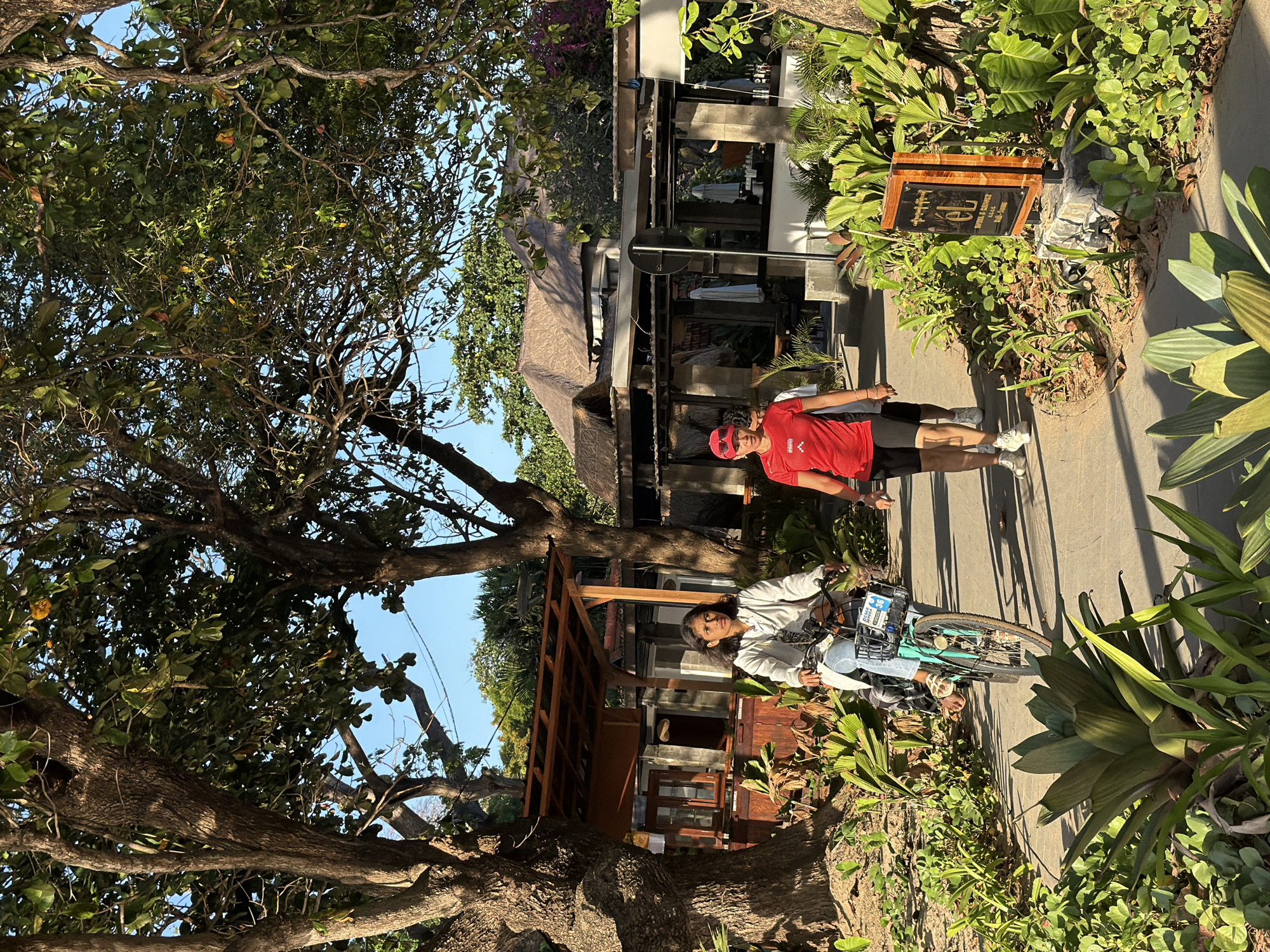 In Denpasar, Indonesia, a young woman rides a teal bicycle with a basket, wearing a light gray top and blue jeans. She is on a paved path, shaded by large trees with lush green leaves. To her right, a woman in a red athletic shirt and black shorts walks towards the viewer, her face partially obscured by sunglasses. Behind them, a resort-like structure with a thatched roof and open-air seating is visible. A wooden sign with "EL HOTEL & RESORT" is positioned to the right of the path, surrounded by tropical plants. The sun casts dappled shadows on the path and foliage.
