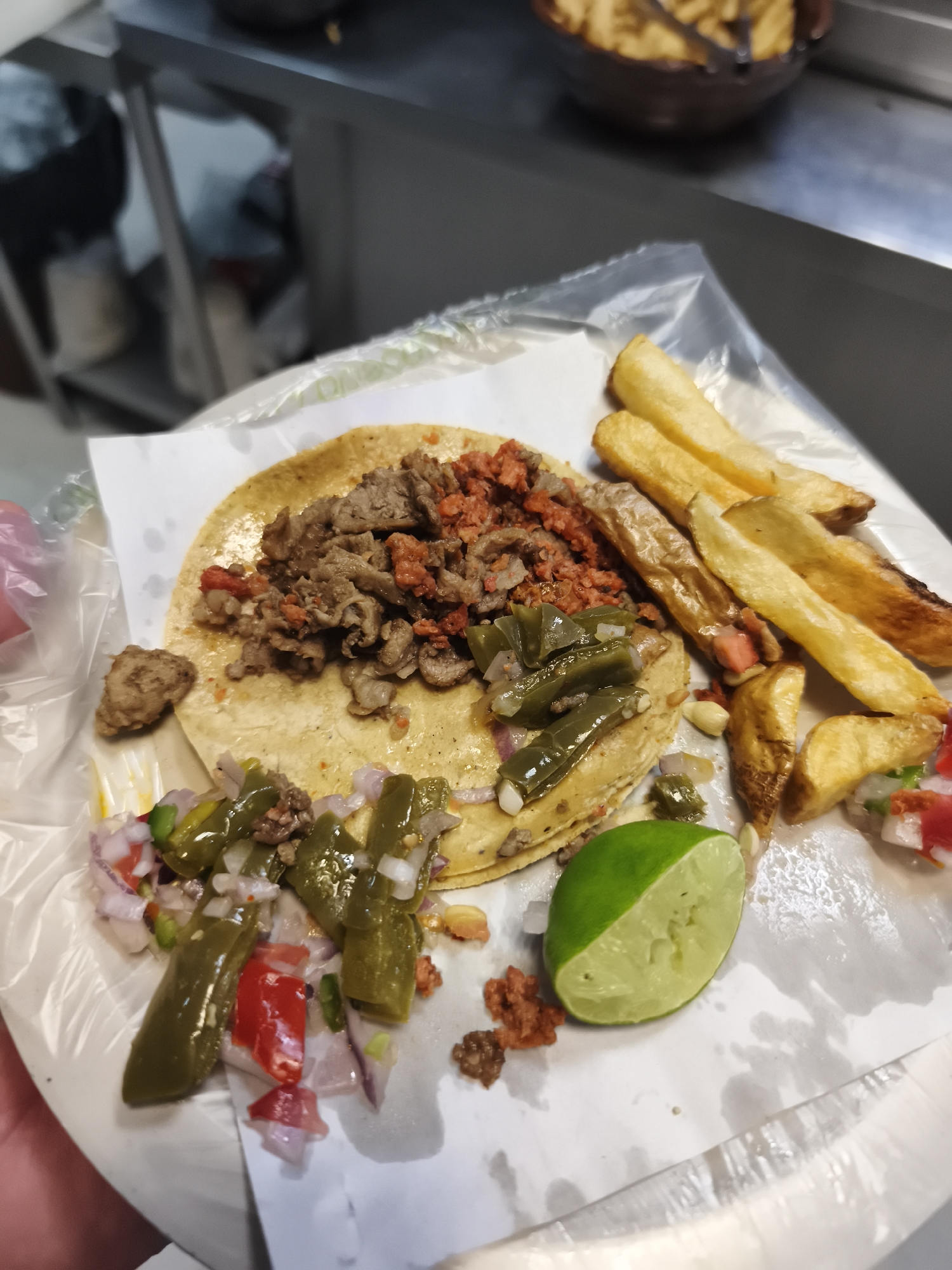 A close-up shot shows a plate of food, likely a taco meal, placed on a surface with what appears to be a metallic backdrop. The plate is a plain white color and sits atop a sheet of clear plastic. The food consists of a tortilla topped with meat, accompanied by what looks like a serving of french fries, diced nopales (cactus), a salsa-like mixture, and a half lime. The meat on the taco appears to be a combination of steak and chorizo. The overall presentation suggests a casual setting, possibly a street food vendor or a small eatery, given the presentation on disposable materials. While the image does not confirm the location, the dish suggests it could potentially be located in Mexico City, Mexico due to the prevalence of tacos and nopales in the cuisine. The environment is indoors, with some blurry background elements suggesting a kitchen or food preparation area. The image captures a still-life moment of a food serving ready to be consumed.