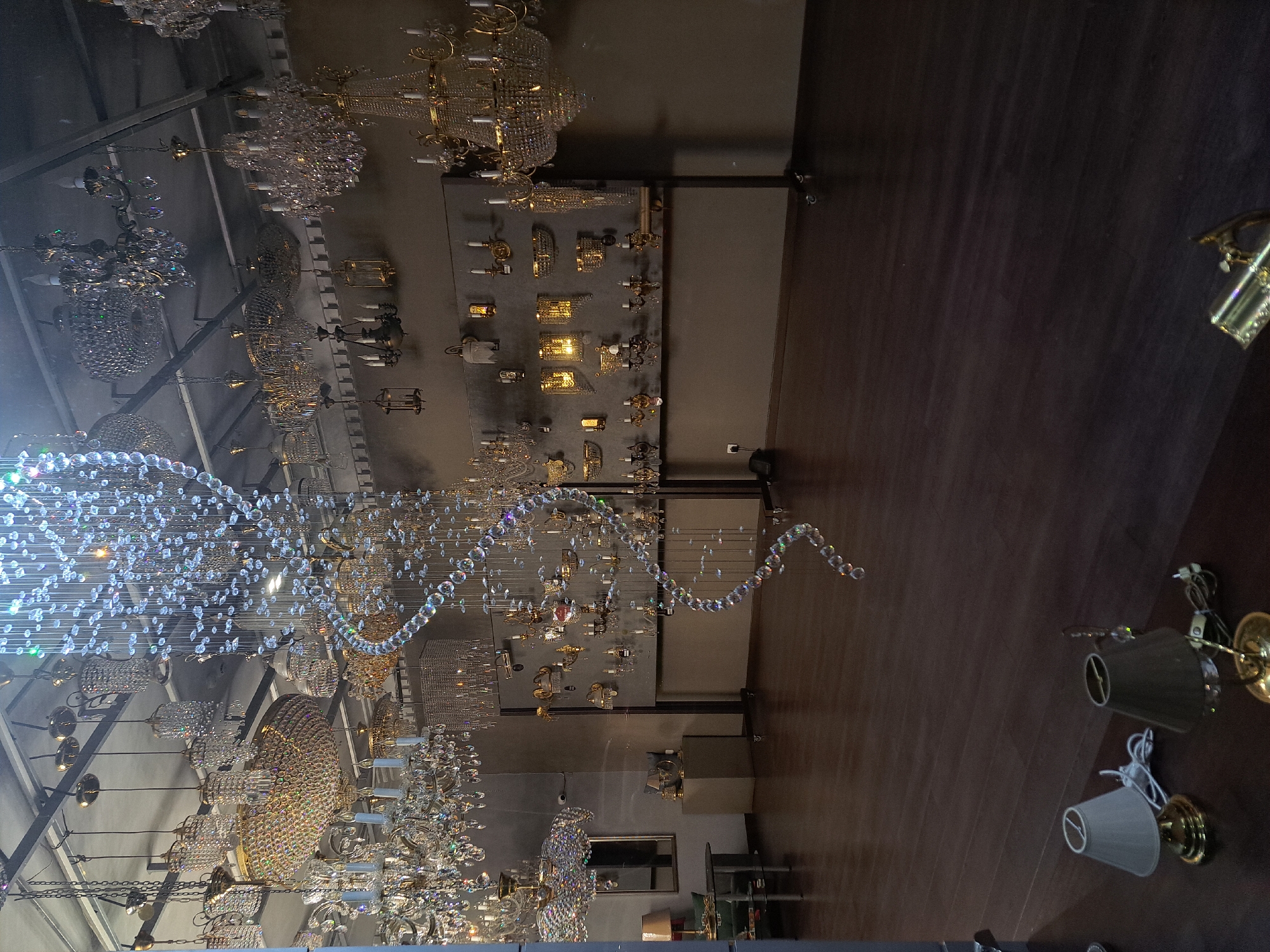 The photograph appears to be taken inside a lighting store, likely in Yerevan, Armenia, given the user's prompt. The main subjects are numerous chandeliers of various sizes and styles, predominately crystal and gold, hanging from the ceiling. Some are displayed on the walls. The ceiling is high and angled. There are examples of wall sconces also visible. The flooring appears to be a dark, polished wood or similar material. No people are visible in the frame. The environment is well-lit.