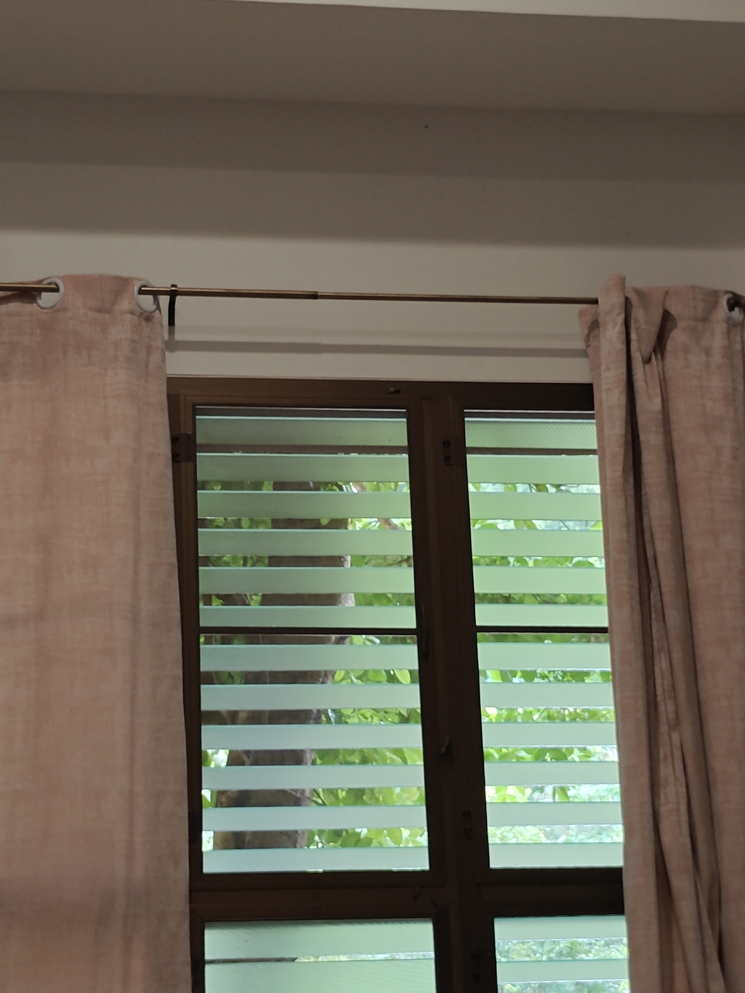 The image shows a close-up view of a window with horizontal blinds, partially obscured by draped curtains on either side. The window frame appears to be made of dark wood. Through the blinds, lush green foliage is visible, suggesting an outdoor environment. The lighting is soft and diffused, creating a serene atmosphere. The location is identified as Mu Si, Thailand, which is known for its mountainous terrain and agricultural landscapes, often featuring fruit orchards and scenic viewpoints.