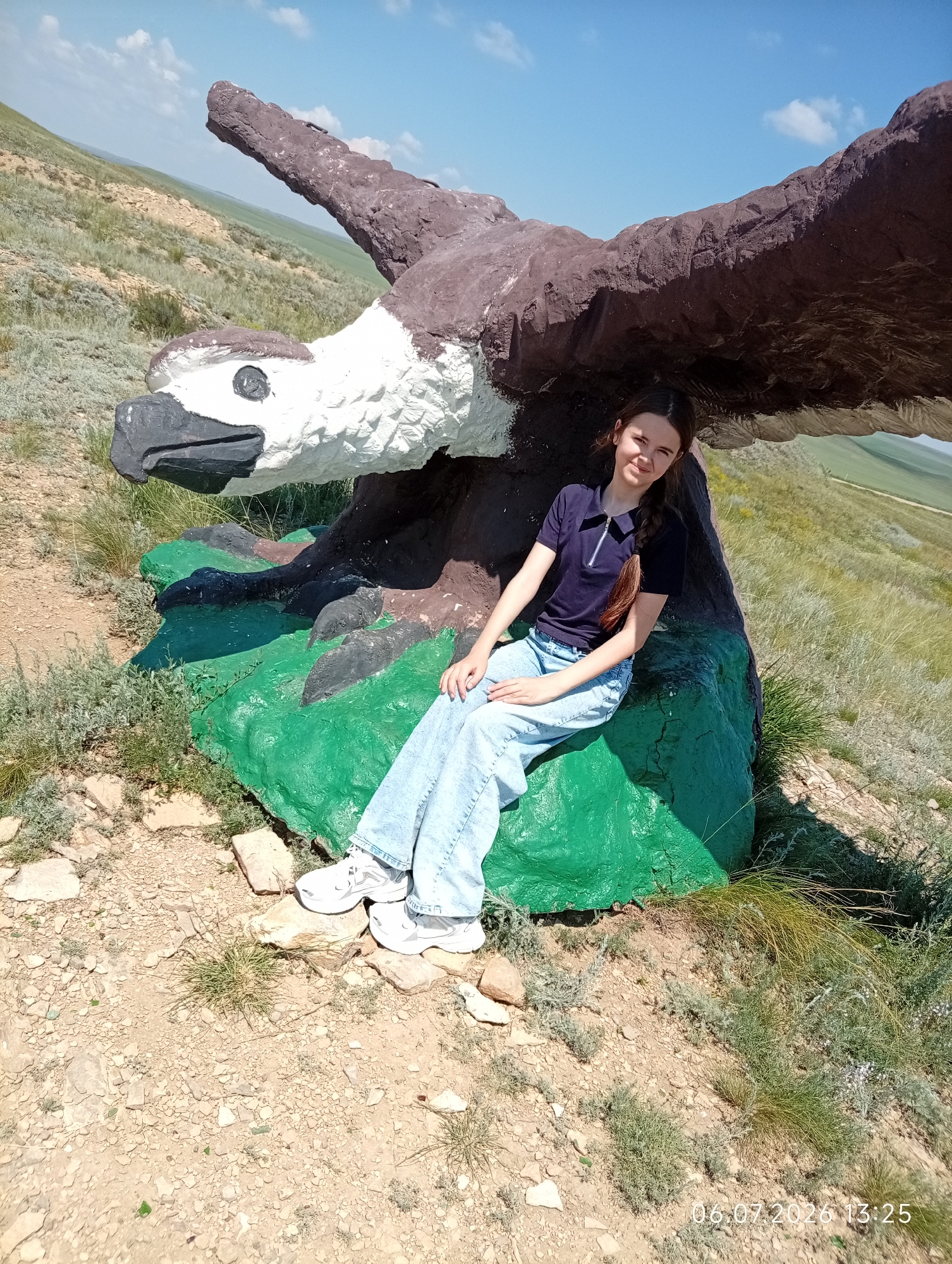 A young woman with dark hair braided to the side is seated on a large, green, rock-like structure painted to resemble the base of a giant eagle sculpture. The sculpture's head, with a white face, black beak, and white-rimmed eye, is visible to the left. The eagle's large, brown, outstretched wings extend over the woman. She is wearing a dark blue polo shirt with a zipper and light blue jeans. Her feet are clad in white sneakers. The scene is set outdoors on a dry, grassy hillside under a clear blue sky with scattered clouds. The ground is covered with dirt, rocks, and sparse vegetation. In the background, rolling hills stretch into the distance. The timestamp on the image indicates it was taken on July 6, 2026, at 13:25. This location is identified as Aleksandrovka, Kazakhstan.